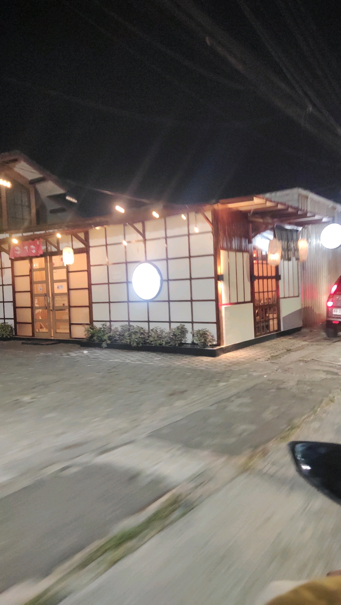 This is a night-time, outdoor, slightly blurred image of a building with Japanese-inspired architecture in Grendeng, Indonesia. The building has a light-colored facade with a grid pattern and wooden accents. There are several windows and doors. A circular illuminated sign is visible on the front of the building. The area in front of the building appears to be a paved lot, possibly a parking area. The sky is dark, and some overhead power lines are visible. The image seems to be taken from a moving vehicle, as indicated by the motion blur in the foreground. A portion of a dark object, likely a side-view mirror of a vehicle, is visible in the lower right corner. A red vehicle is parked on the right side of the building. Small plants are arranged in front of the building's facade.