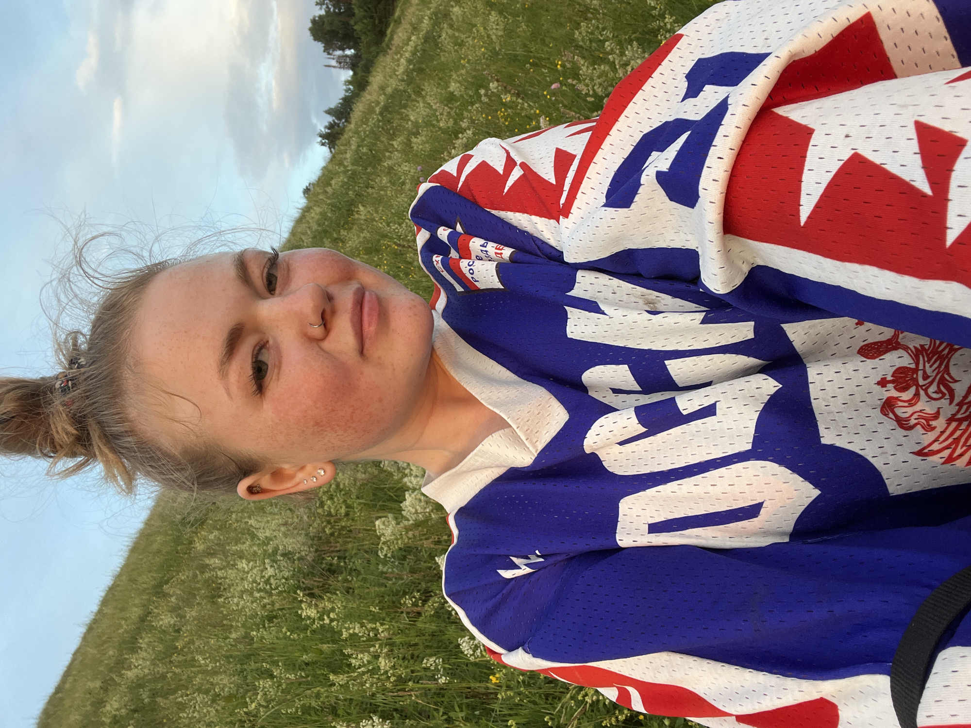 A young woman with blonde hair in a messy bun and freckles across her nose and cheeks is pictured from the chest up, wearing a blue, white, and red hockey jersey. She has a nose ring and multiple earrings. The woman is outdoors, seated in a grassy, hilly area with scattered wildflowers. The sky is visible in the background, with clouds and a hint of trees on the horizon. The setting appears to be a natural landscape on the outskirts of Olkhovka, Russia.
