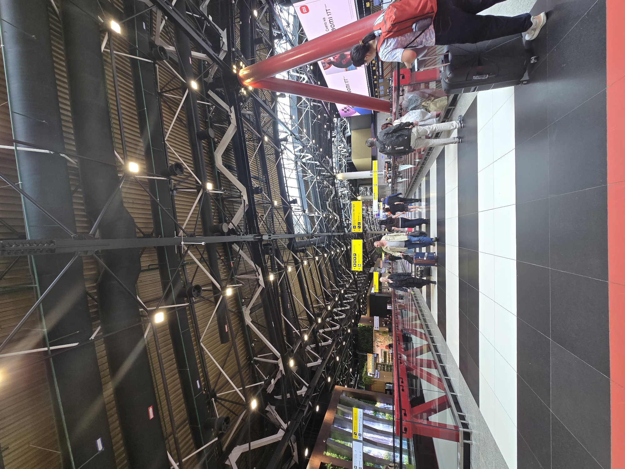 The image depicts a large, modern indoor space, likely an airport terminal or a major transportation hub, consistent with its location in Khimki, Russia. The perspective looks down a long, high-ceilinged hall from a slightly elevated vantage point.
The overhead structure is a prominent feature, showcasing a complex industrial design with a vast network of black metal trusses, white support beams, and large black cylindrical ductwork. Numerous bright square LED light fixtures are embedded within this structure, illuminating the space. Striking red, tubular structural columns punctuate the ceiling's vastness, providing visual anchors. Some natural light seems to enter from windows or skylights along the upper section of the ceiling.
The floor beneath is distinctive, featuring a bold pattern of alternating black and white stripes that stretch into the distance, guiding the eye. Red borders or pathways run along the sides of this striped area.
Several individuals and small groups of people are scattered across the floor, all appearing to be travelers. Many are equipped with luggage, including suitcases and backpacks. They are engaged in activities typical of a travel hub, such as walking, standing, or waiting. One person in the foreground, wearing a red backpack, is seen looking at a mobile phone while standing next to a black suitcase.
Visible text includes a large digital advertising screen in the background displaying "Xiaomi 11T" and other text, likely in Russian. Additionally, several smaller yellow rectangular signs with symbols and foreign text are suspended from the ceiling, probably providing directional information or gate numbers. The time of day appears to be daytime, and the overall atmosphere is bright and bustling, characteristic of a busy travel environment.