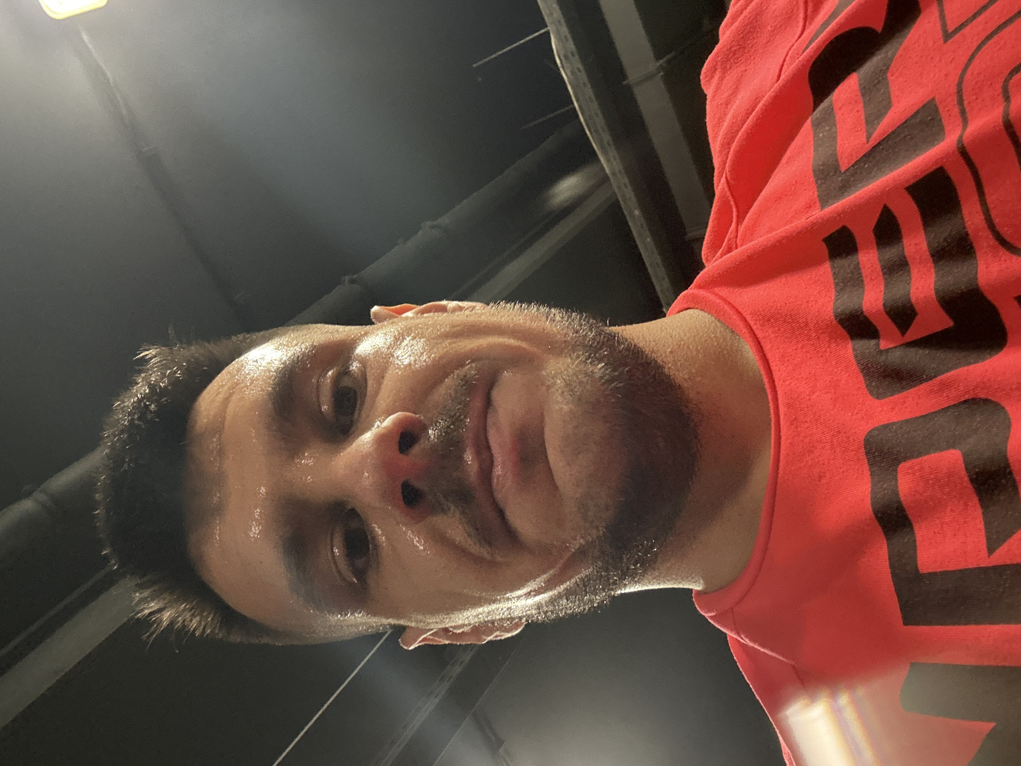 A close-up, low-angle shot depicts a sweaty man in a red t-shirt with black lettering. The man has dark hair, a beard, and appears to be perspiring heavily. The background consists of a dark ceiling with metal beams and possibly a light fixture. The image provides no information to indicate the location in Sofia, Bulgaria or any details beyond the immediate scene.