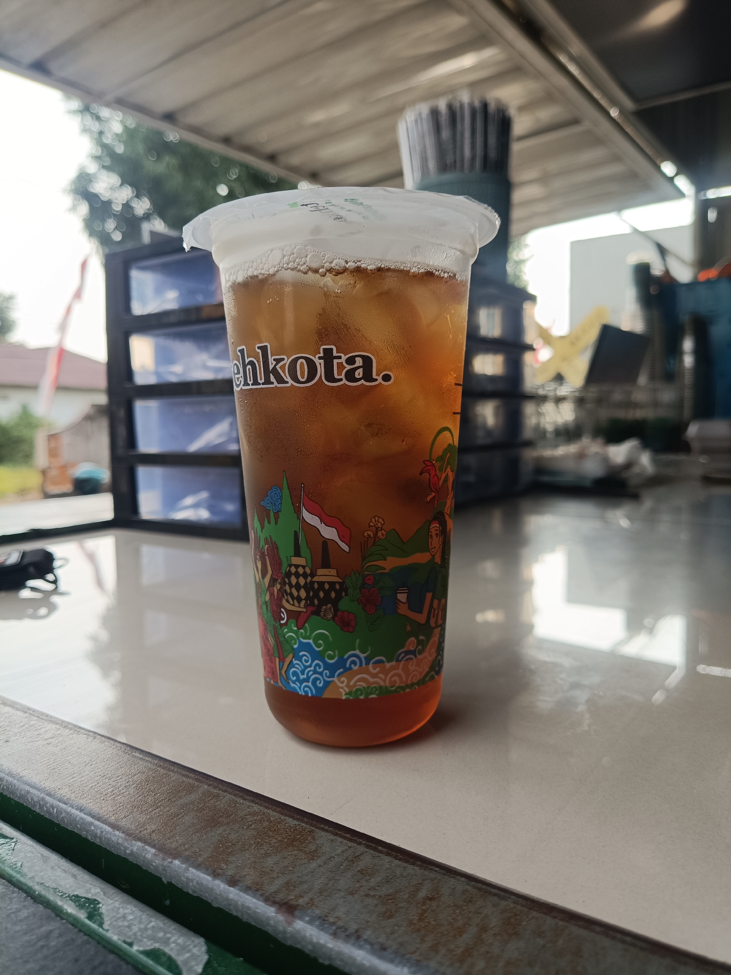 A close-up, eye-level shot shows a clear plastic cup filled with iced tea. The cup has a white plastic lid with condensation on the outside. The tea is a deep amber color, and ice cubes are visible within. The cup's exterior is decorated with colorful graphics depicting a village scene, including a flag of Indonesia, buildings, people, and nature. The brand name "ehkota." is visible in white text on the upper portion of the cup. The cup sits on a reflective, light-colored surface, possibly a table or counter, which shows reflections of the cup and the surrounding environment. In the background, out of focus, are a dark cabinet with blue drawers, some greenery, and structures that suggest an outdoor or semi-outdoor setting, possibly a food stall or cafe. The scene is well-lit, with natural light contributing to the overall brightness.
The location is identified as Jember, Indonesia, suggesting this scene is part of the local cityscape or a common vendor's setup in that region. The overall impression is that of a refreshing beverage being enjoyed in a casual setting within Jember.