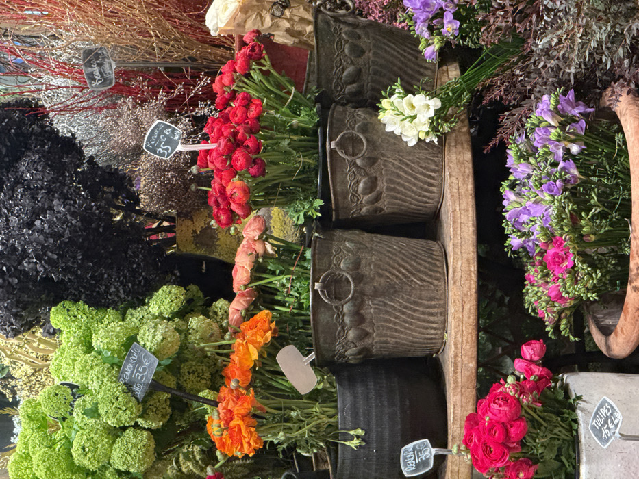 An array of flowers and foliage fills the image. Red, orange, and blush-toned ranunculus stand next to green hydrangeas and black decorative branches. Purple and pink freesia are arranged near brown, textured metal buckets. Small tags indicate prices. The scene resembles a flower shop display, showcasing a variety of colors and textures.