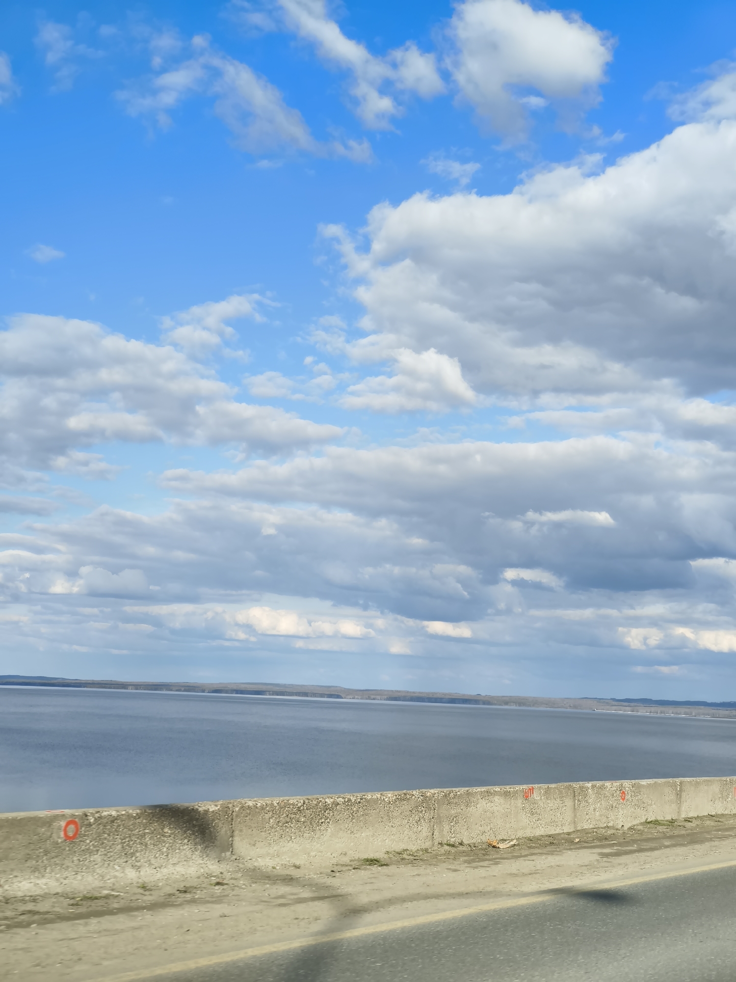 The image displays a wide landscape view from a roadside or bridge overlooking a vast body of water, likely a river or reservoir, consistent with the location context of Noviy, Russia. The scene is captured during the daytime under a partly cloudy sky, with patches of bright blue visible amidst numerous white and grey cumulus clouds stretching across the upper two-thirds of the frame.
In the foreground, a weathered concrete barrier or railing runs horizontally, separating the viewer's immediate perspective from the expansive water. Below this barrier, a section of an asphalt road or shoulder is visible, featuring a faint yellow line and subtle reflections. The large body of water occupies the middle ground, appearing calm and reflecting the sky's light, with a faint, dark landmass or distant shoreline visible on the far horizon. No people or specific activities are present, giving the scene a serene and expansive feel.
Notable details include several red markings painted onto the concrete barrier. On the far left, a distinct red circle with a central dot is visible. Further to the right, additional red marks, some appearing to be numbers such as "107" and "3," are discernible, suggesting they might be survey points, distance markers, or other annotations on the structure. The overall impression is one of an open landscape viewed from an elevated, man-made vantage point.