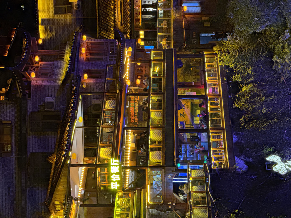 The image displays a multi-level commercial complex built into a natural rock face or cliffside at night in Furong, China. The structures feature traditional Chinese architectural elements, including dark brick walls, tiled roofs with upturned eaves, and wooden balconies. The entire scene is brightly illuminated by numerous warm yellow lights, string lights outlining architectural features, and hanging lantern-style fixtures. A prominent yellow sign on an upper level reads "平价餐厅" (Affordable Restaurant).
Multiple stories contain both enclosed interior spaces with large windows and open-air balconies. Numerous individuals, estimated at 30-40, are visible across the various levels, primarily seated at tables within these establishments. They appear to be dining or socializing. On a lower level, a bar counter with bottles and glasses is visible, with several patrons seated nearby. Another partial sign on a lower level reads "不...饭店".
The complex is constructed above a steep, dark natural slope covered with dense, dark green foliage, including vines and bushes. A glowing, abstract, light-colored neon-like art piece is affixed to the foliage on the lower left. The sky is dark, indicating a nighttime setting. A lone individual in dark attire stands on an outdoor walkway on the lower right side of the image. Air conditioning units are mounted on the exterior brick walls.