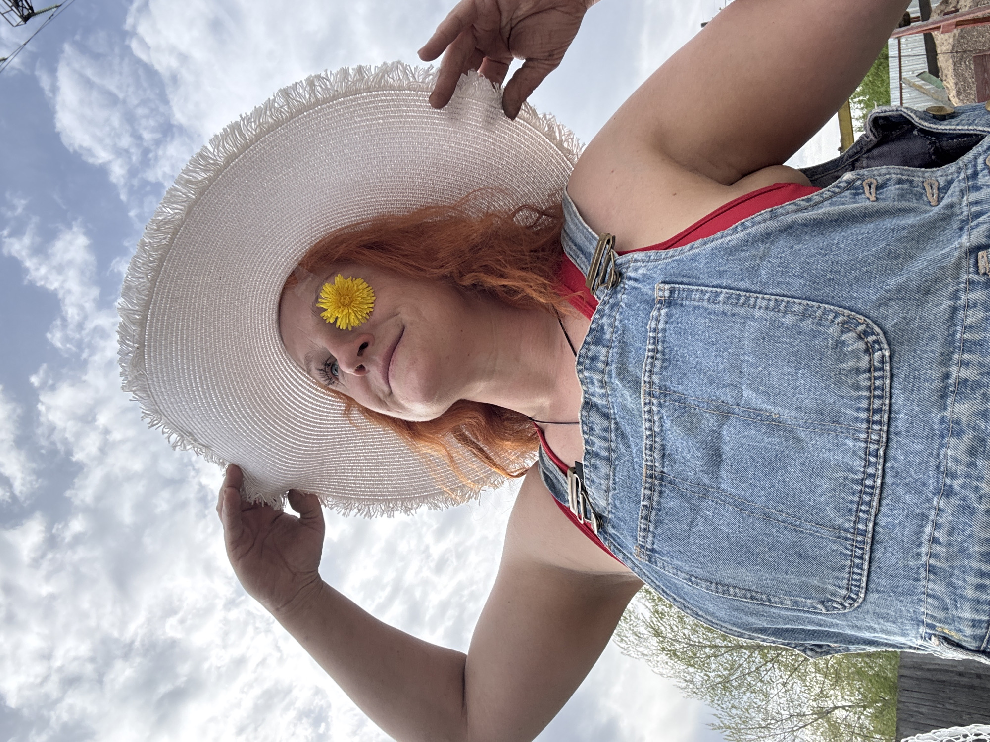 A woman is captured from a low angle, looking upwards. She is wearing denim overalls over a red top and a wide-brimmed straw hat adorned with a yellow flower positioned near her eye. The sky, scattered with clouds, serves as the backdrop. The location is Chaikovsky, Russia.
