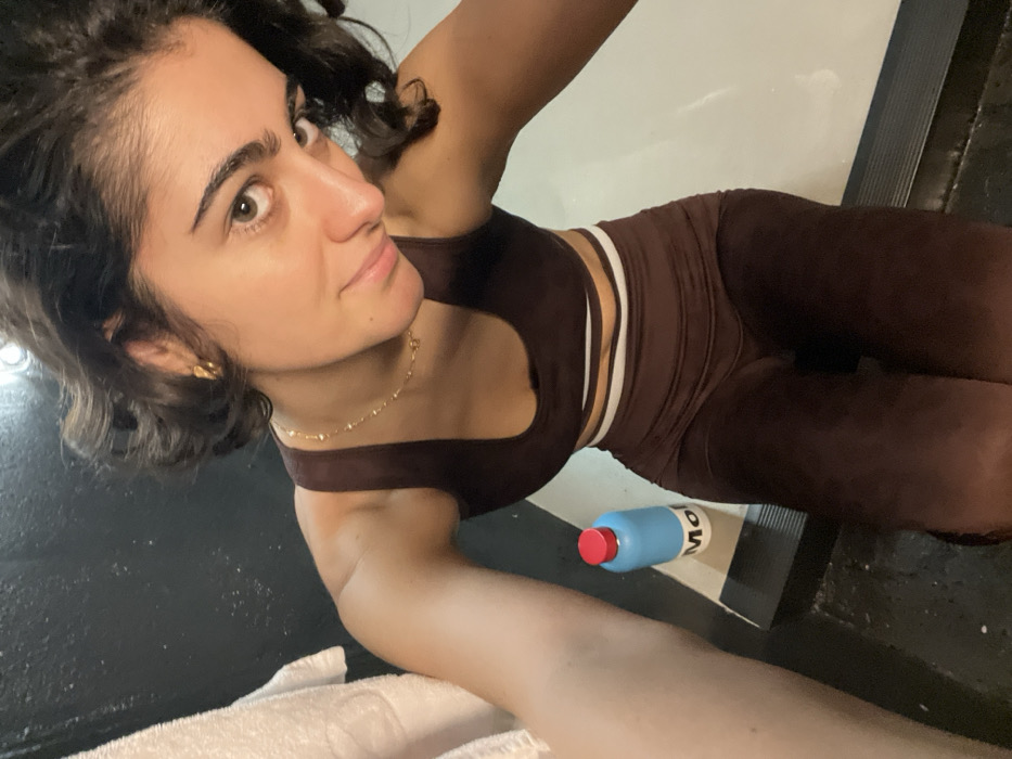 A woman wearing a brown sports bra and matching leggings is angled towards the camera. Her hair is short and curly. She's wearing a gold necklace and earrings. A blue bottle with a red cap sits near her waist on a black and white striped mat. A white towel is visible in the lower left corner. The background includes black flooring and a white wall. The lighting is bright.