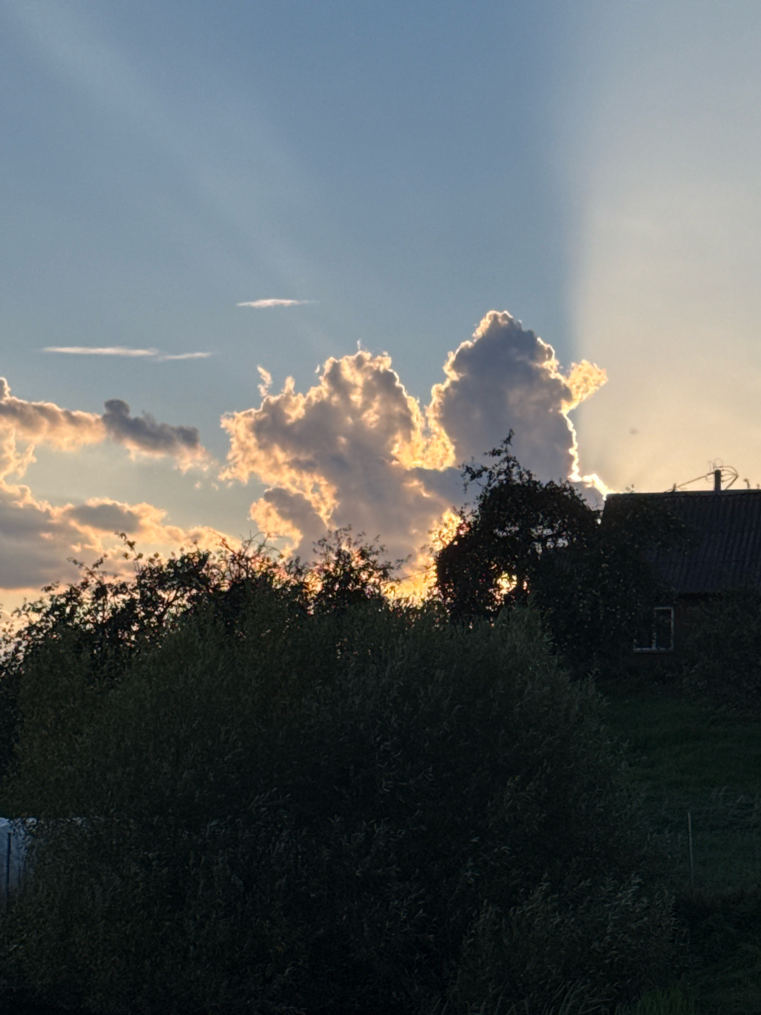 A photograph taken at dusk in Galinė, Lithuania, shows a natural landscape with a partially obscured residential building. The sky is dominated by the late afternoon or early evening sun, casting dramatic rays through the clouds. The clouds themselves are illuminated from behind by the setting sun, creating a bright, glowing effect along their edges and on their undersides. In the foreground, dense foliage of trees and bushes obscures the lower portion of the image. To the right, a portion of a dark-colored, sloped roof of a building is visible, with a small window or opening near its peak. The setting sun creates a distinct line between the brightly lit clouds and the shadowed area of the sky, suggesting the sun is near the horizon. The overall scene depicts a serene, rural twilight atmosphere.
