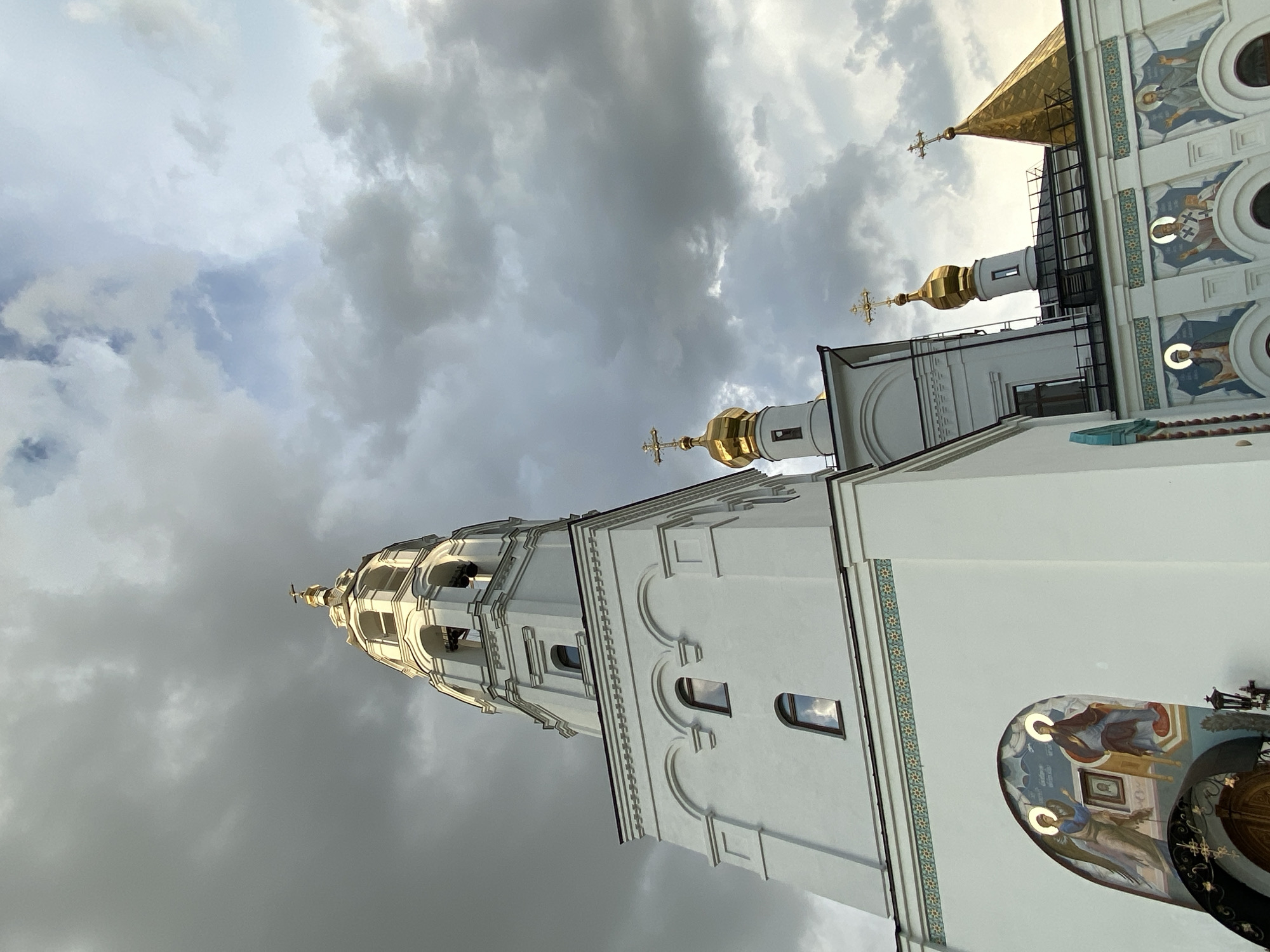 The image shows a low-angle view of a white church with golden domes against a cloudy sky. The church features a tall bell tower with arched openings and a smaller tower topped with a golden dome and a cross. The exterior walls are adorned with frescoes depicting religious figures. The overall scene suggests a religious site located in Yekaterinburg, Russia, likely on a day with overcast weather. There are no people visible in the image.