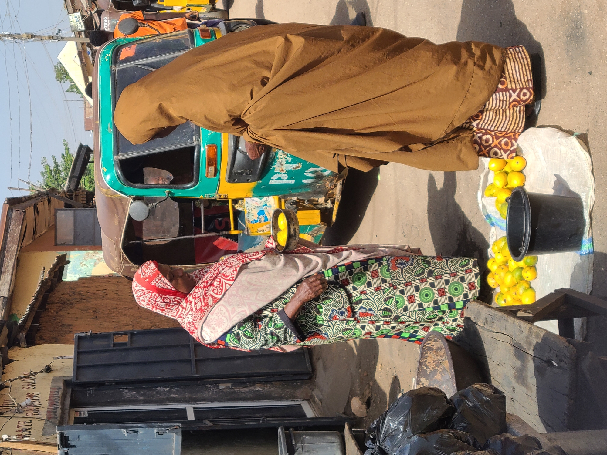 The image shows an outdoor scene in Jos, Nigeria. A woman in colorful patterned clothing and a red headscarf is standing near a wooden bench and a black trash bag. Next to her, a white bag filled with yellow fruits (possibly mangoes) sits next to a black bucket. A second person draped in brown fabric is partially visible near a yellow and green auto-rickshaw. The background includes buildings with corrugated metal roofs and signs. The scene is lit by sunlight.