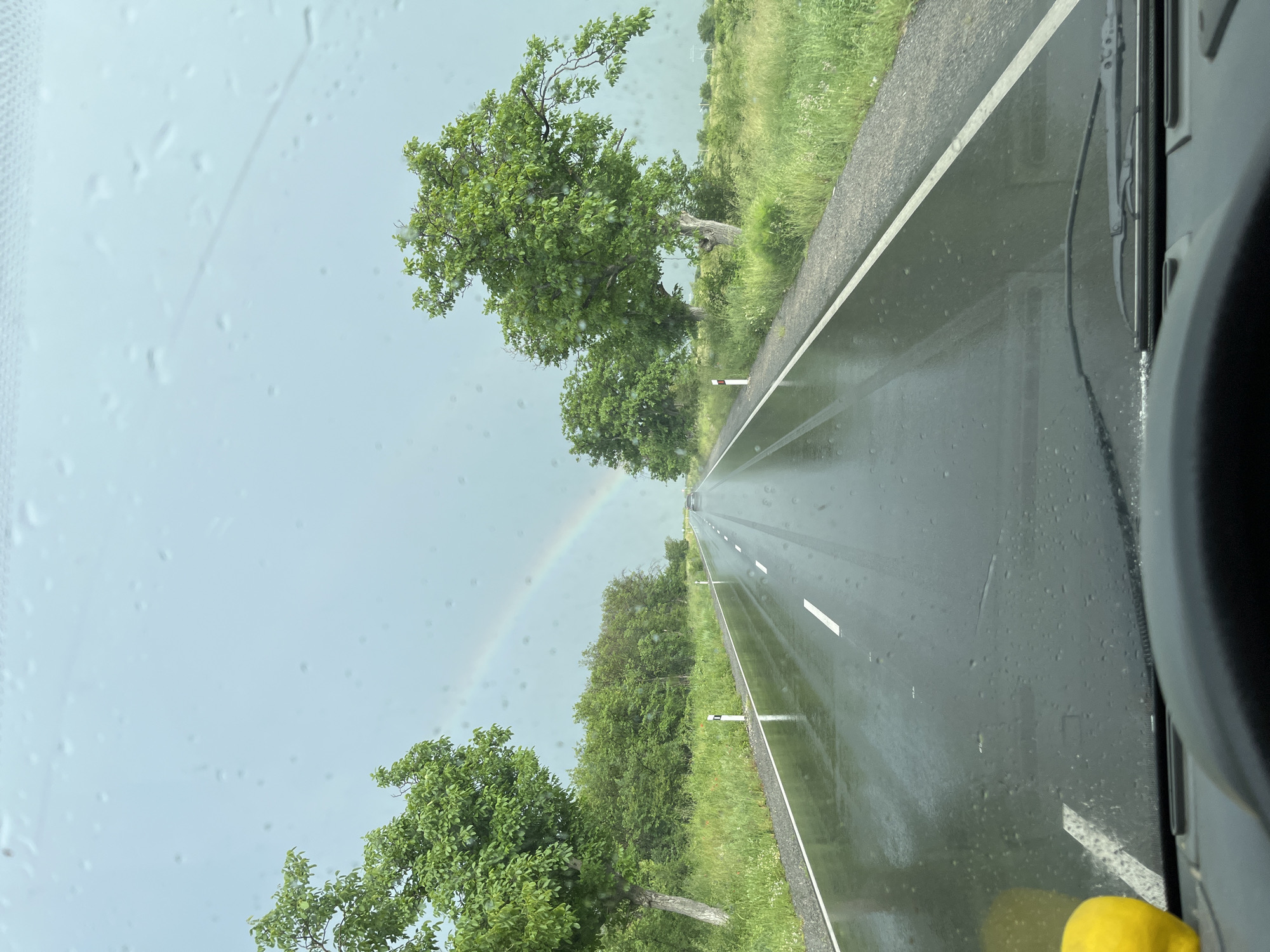 The image shows a view from inside a vehicle, looking out onto a wet asphalt road stretching into the distance. The perspective is slightly angled. There are white lane markings on the road. Trees line both sides of the road. A rainbow is visible in the sky. The image was taken while driving through the city of Pevtsite, Bulgaria.