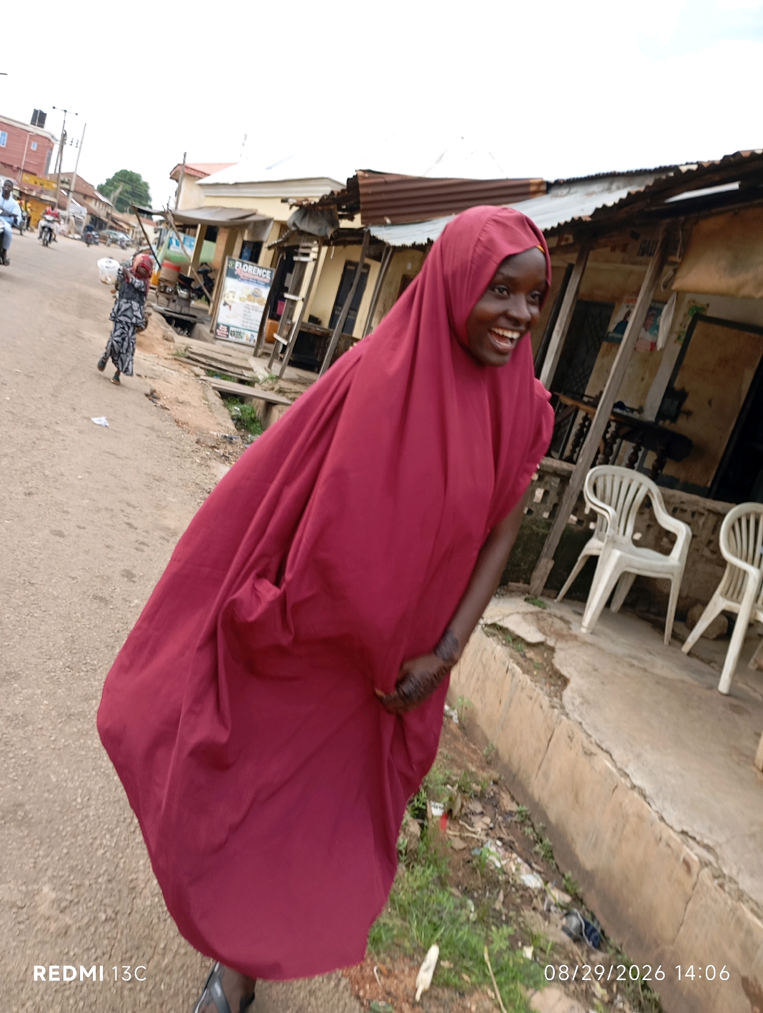 A young African female, wearing a maroon hijab and dress, smiles widely while standing on a dusty, unpaved road. Her right hand, adorned with henna, is raised to her face. In the background, a street scene unfolds with several shops, some with corrugated iron roofs. Two white plastic chairs are visible outside one of the shops. A young child in a patterned outfit walks away from the viewer down the road, and a motorcycle is seen in the distance. The scene appears to be in Shendam, Nigeria, as indicated by the timestamp and potentially the architecture and local attire.