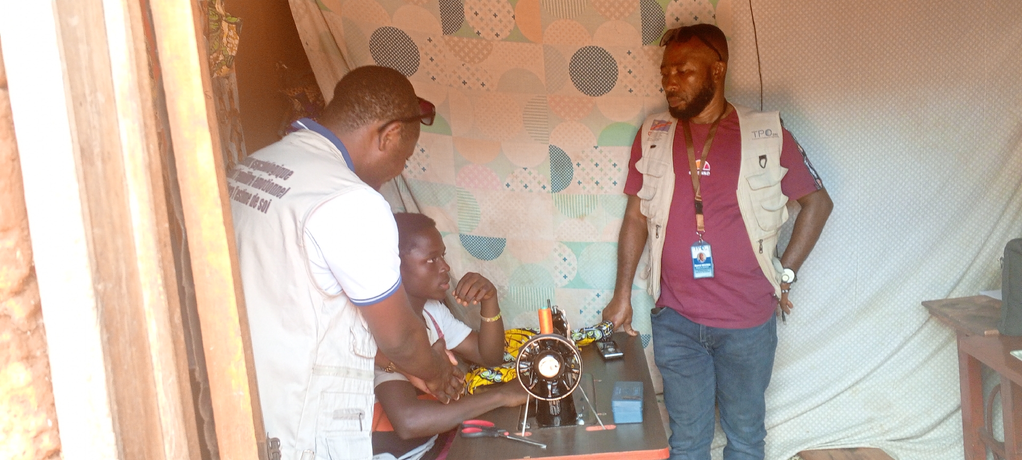 This image captures three men in an indoor or semi-enclosed setting, likely a workshop or training area, in Ngalula, Democratic Republic of the Congo. The background is a patterned fabric, and a wooden structure is visible on the left.
In the foreground, a young man sits at a black manual sewing machine, focusing intently. Colorful fabric, scissors, and pliers are on the table next to the machine. Another man stands directly behind him, leaning over as if providing guidance or instruction. This second man wears a white short-sleeved shirt with blue trim and a light-colored vest. Text visible on his shirt, partly obscured, includes French words such as "...psychologique" (psychological), "...émotionnel" (emotional), and "...estime de soi" (self-esteem). He also wears sunglasses pushed up onto his head.
To the right, a third man stands, observing the scene. He is wearing a maroon t-shirt, blue jeans, and a light-colored multi-pocket vest. An ID badge hangs around his neck, displaying the acronym "TPC" and a small photograph. The vest itself also has "TPO" printed on one pocket. Sunglasses are also on his head. A logo on his t-shirt appears to be "TATA".
The activity depicted is likely a vocational training session or mentorship in sewing, with the men demonstrating or learning practical skills. The setting is simple, and the bright lighting suggests it is daytime. All three individuals appear focused and engaged.