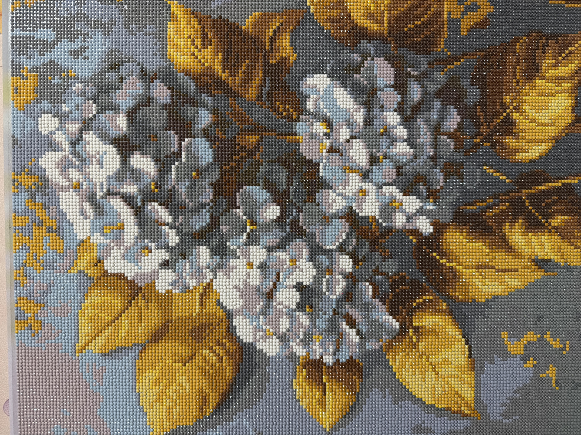 The image is a close-up, high-angle, full-frame shot of a diamond painting depicting a floral arrangement of hydrangeas and golden leaves against a mottled grey and blue background. The painting is made up of thousands of small, faceted resin beads, each a distinct color, creating a mosaic-like effect. The hydrangeas are primarily rendered in shades of white, light blue, and pale pink, with small yellow-orange centers. The leaves are a vibrant golden yellow and various shades of brown, with visible veins and textured surfaces. The background is a blend of grey, light blue, and pale pink, with some darker grey areas that suggest shadow. The overall impression is one of a highly detailed and colorful artwork, likely a craft project. There is no indication of a specific location in Kyiv, Ukraine, or any people or actions depicted.