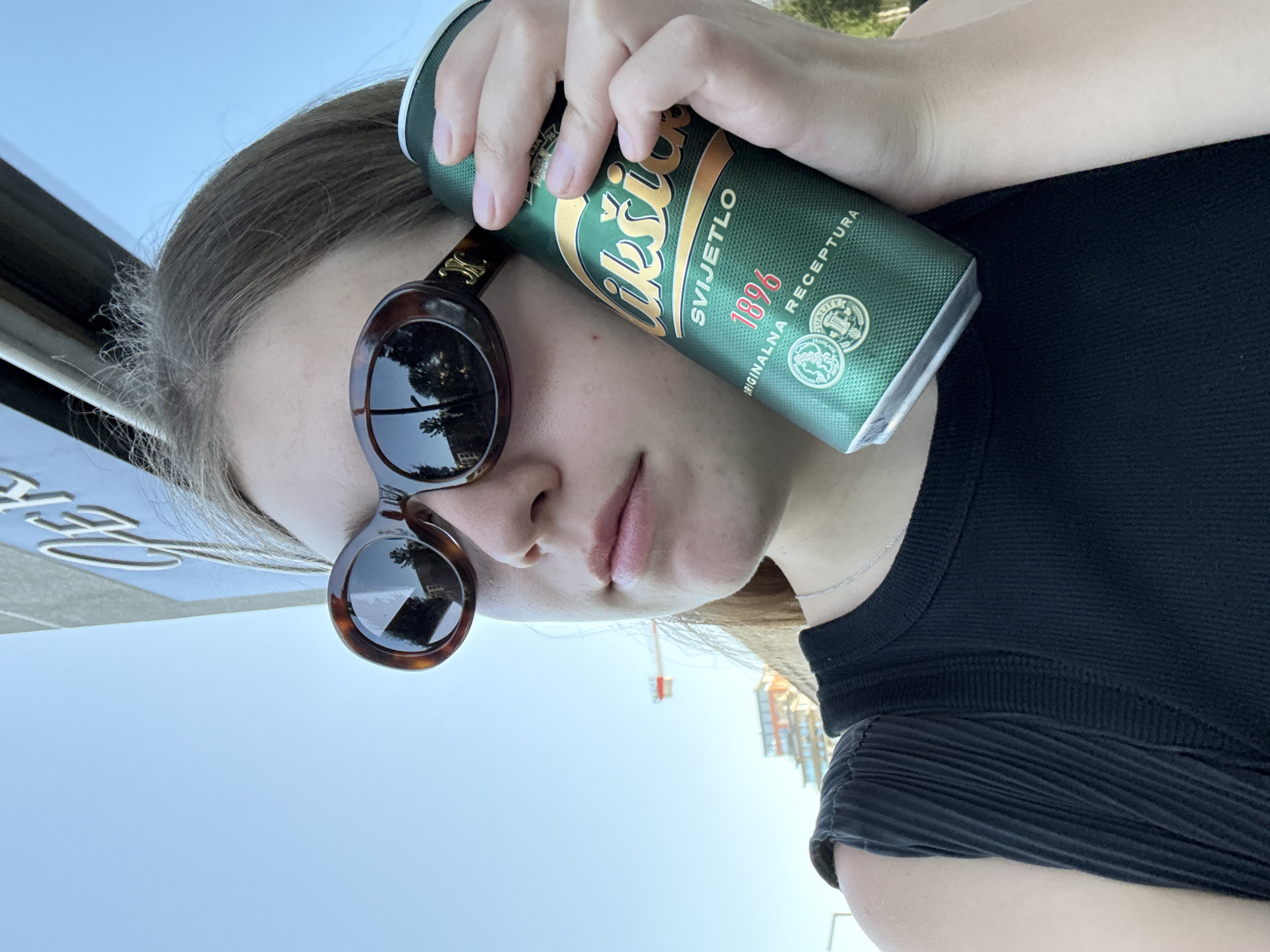 A person wearing sunglasses and a dark-colored shirt holds a green and gold can of "Nikšićko Svijetlo 1896" beer. The background includes a bright blue sky, a partially visible building or structure with text, and a distant view of buildings and a crane. The photograph appears to be taken outdoors during daylight hours. The location is likely Belgrade, Serbia, as Nikšićko beer is a popular brand in the region.