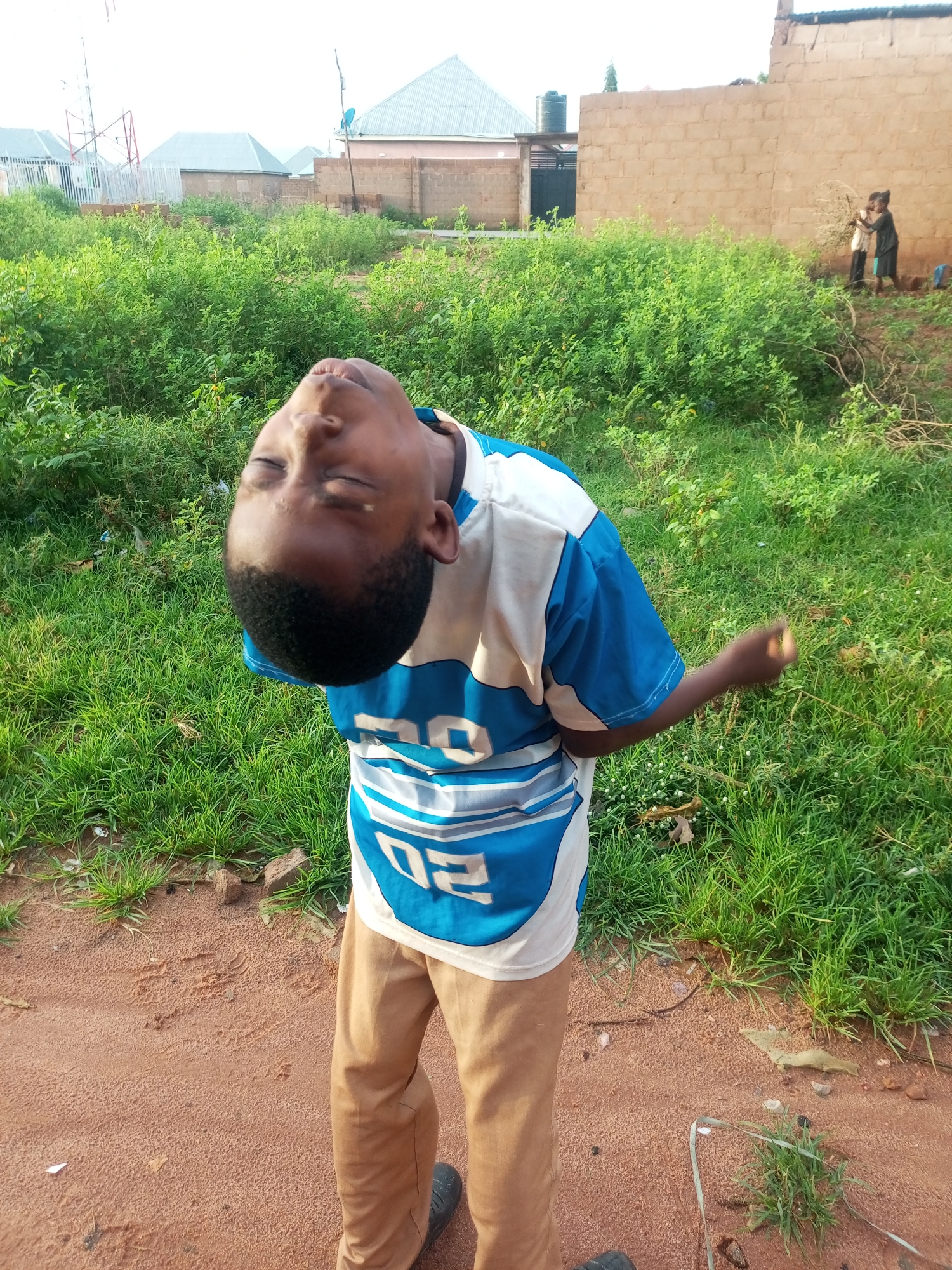 In an outdoor scene in Tempure, Nigeria, a young boy is captured mid-motion, his head tilted back with his eyes closed, and his body appearing to be in a dance or celebratory pose. He is wearing a blue and white t-shirt with a horizontal striped design and beige pants. The background reveals a residential area with low-rise buildings, some with corrugated metal roofs, and a patch of lush green vegetation. Two other figures, possibly children, are visible in the distance near a brick wall. The ground is a mix of reddish-brown dirt and patches of grass.