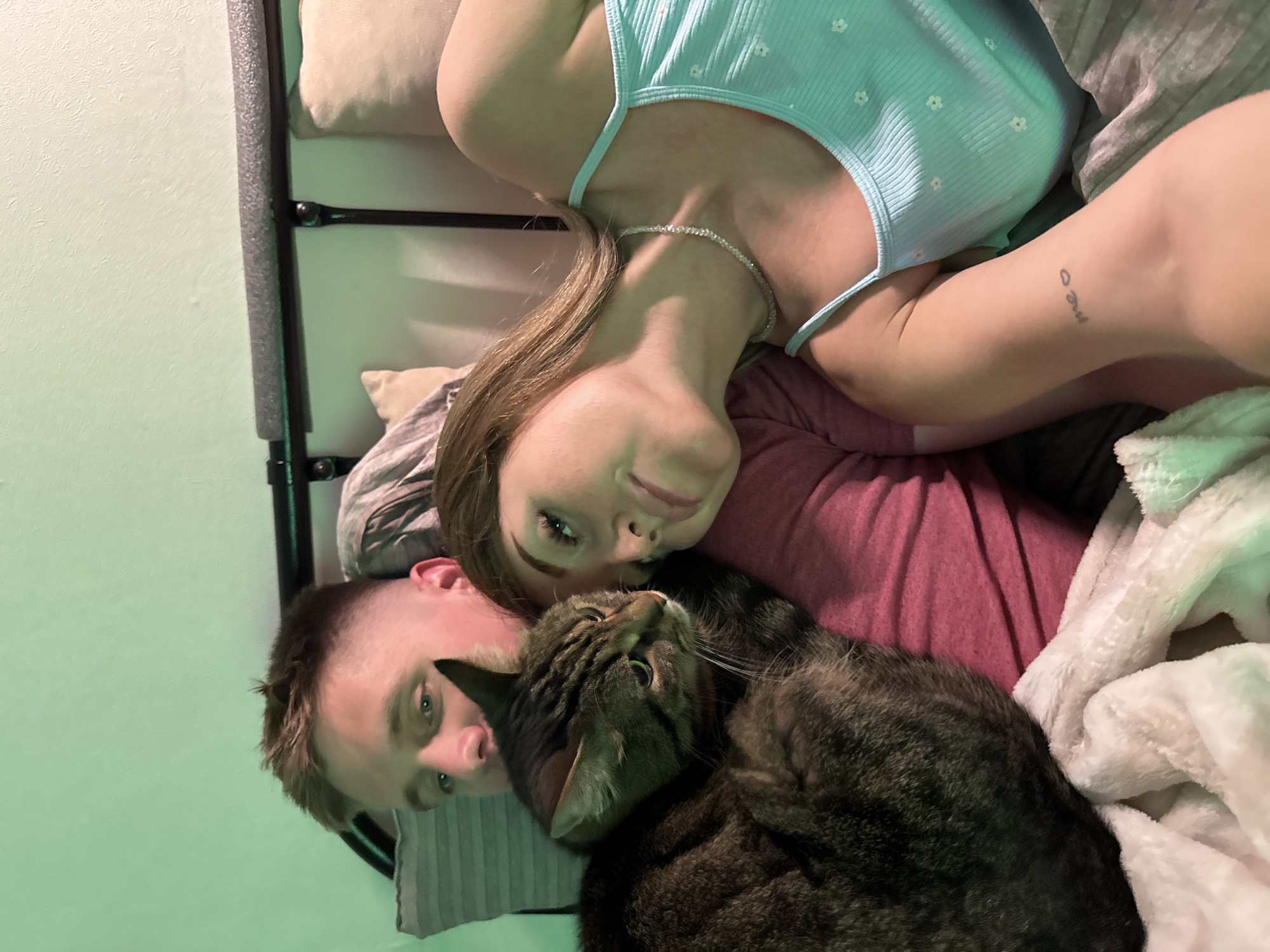 An overhead, close-up shot captures a man and a woman lying in bed with a tabby cat nestled between them. The man's face is visible in the upper left, his eyes open and looking towards the camera. The woman's face is to the right of the man's, with her eyes closed and her head resting on his shoulder. The cat is curled up in the man's arms, its head turned away from the camera, looking to the left. A green light illuminates the scene from the left. The bed has a black metal headboard and is covered with pillows and blankets. The woman is wearing a light blue tank top with a floral pattern and a silver necklace. The man is wearing a dark pink t-shirt. The cat is a domestic short-haired tabby with brown and black stripes. There is a tattoo on the woman's left arm that reads "me o". The image is likely taken indoors. The city of Chaikovsky, Russia, is not depicted or referenced in the image.