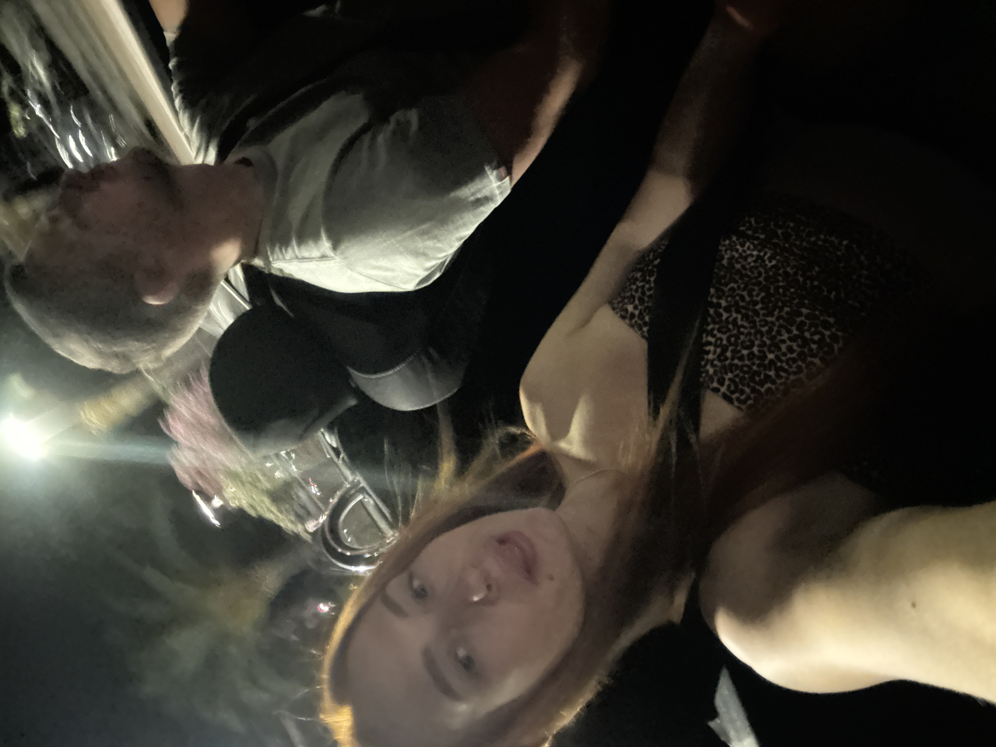 In this media file, two individuals are captured in a close-up shot. The person in the foreground, appearing to be a woman, has light-colored skin and long, reddish-brown hair. She is wearing a leopard-print top. Her face is turned towards the camera, with her mouth slightly open. The second person, likely a man, is partially visible behind her, with light-colored hair and fair skin. He is wearing a light-colored t-shirt.
The setting appears to be indoors or in a dimly lit environment, possibly at night, given the low light conditions and the presence of a bright light source on the left side of the frame, which creates reflections. The overall tone of the image is intimate and casual, suggesting a personal moment between the two individuals. There are no discernible objects or location cues that definitively place this in Budva, Montenegro. The image focuses on the faces and upper bodies of the subjects, with a shallow depth of field that blurs the background. The emotion conveyed is ambiguous due to the low lighting and close cropping, but the overall impression is one of relaxed companionship.