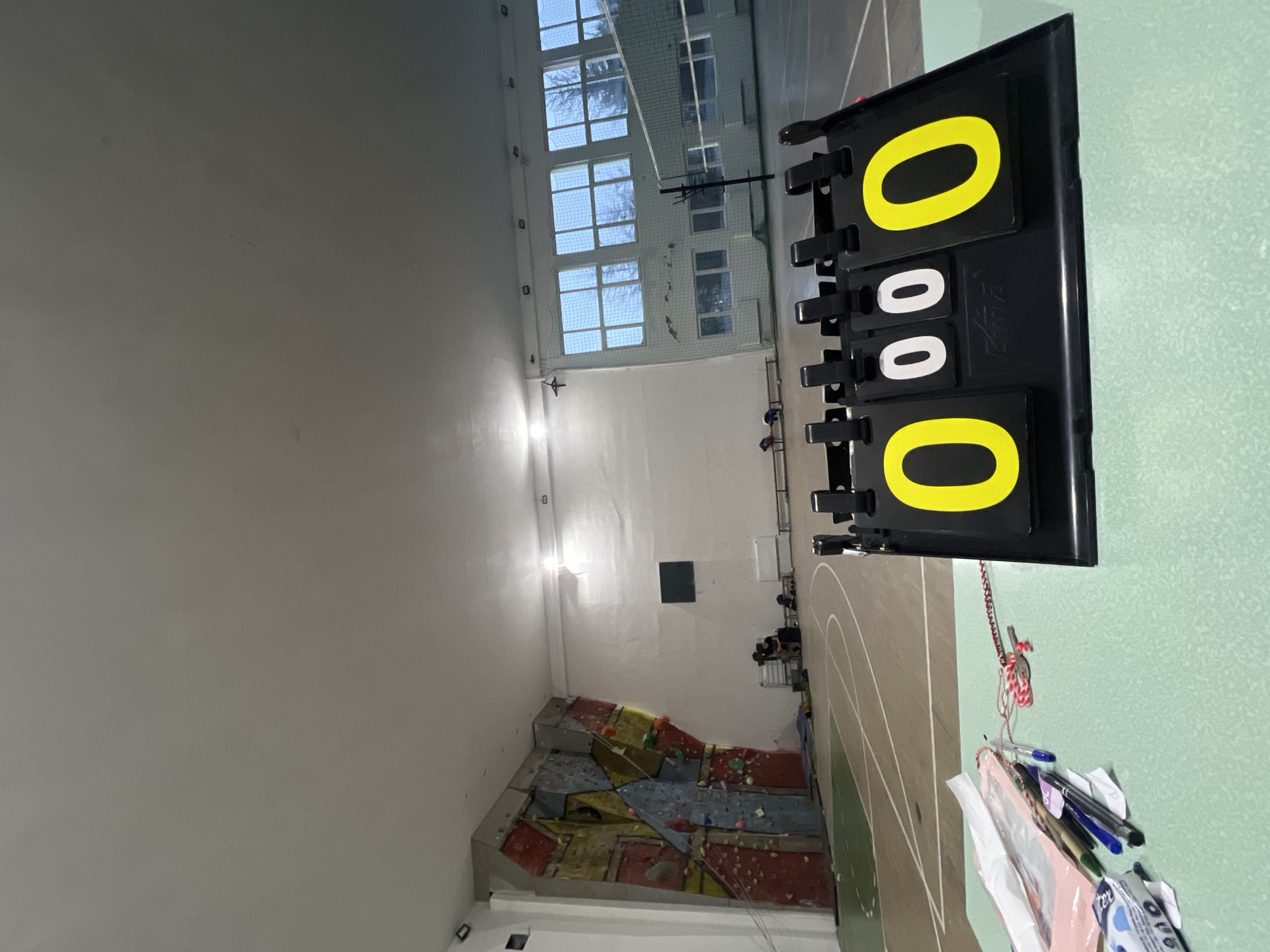 The image depicts the interior of a gymnasium in Elena, Bulgaria. A scoreboard displays the score "000 - 000". A climbing wall is visible in the background. There are individuals situated in the gymnasium, although the image does not allow for precise details about them. The gymnasium has large windows. A climbing net is visible in the distance.