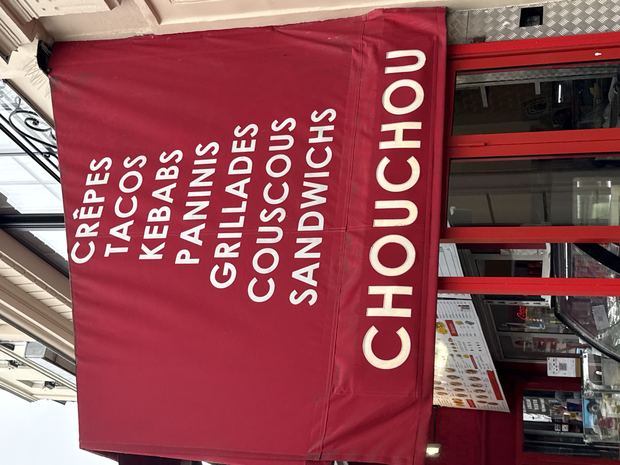 A street-level view of a maroon awning with white lettering, likely in Paris, France, given the food items listed and the architectural style. The awning advertises "CRÊPES," "TACOS," "KEBABS," "PANINIS," "GRILLADES," "COUSCOUS," "SANDWICHES," and "CHOUCHOU." The awning is attached to a building with a red frame around a window. A menu board is visible behind the window. Parts of other buildings and street structures are partially seen in the periphery.