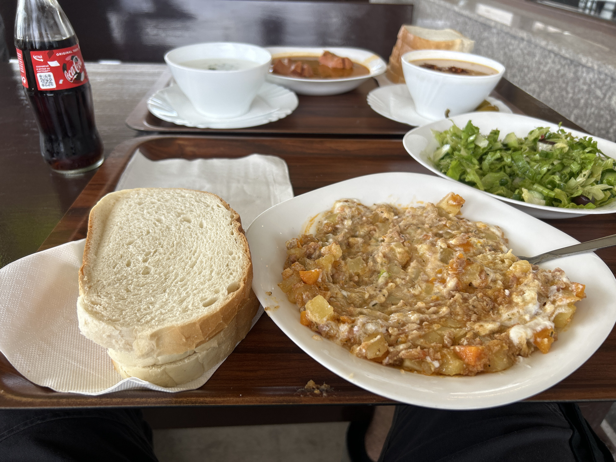 A tray of food is presented in the city of Popovitsa, Bulgaria, featuring a Coca-Cola bottle, a stack of white bread slices on a napkin, a white plate filled with a hearty stew of meat, potatoes, and carrots, a side salad in a white bowl, and two small white bowls containing soup. The food is arranged on a dark wooden tray, which rests on a darker surface. The background shows a blurred environment, possibly a restaurant or dining area, with hints of other tables and seating.
