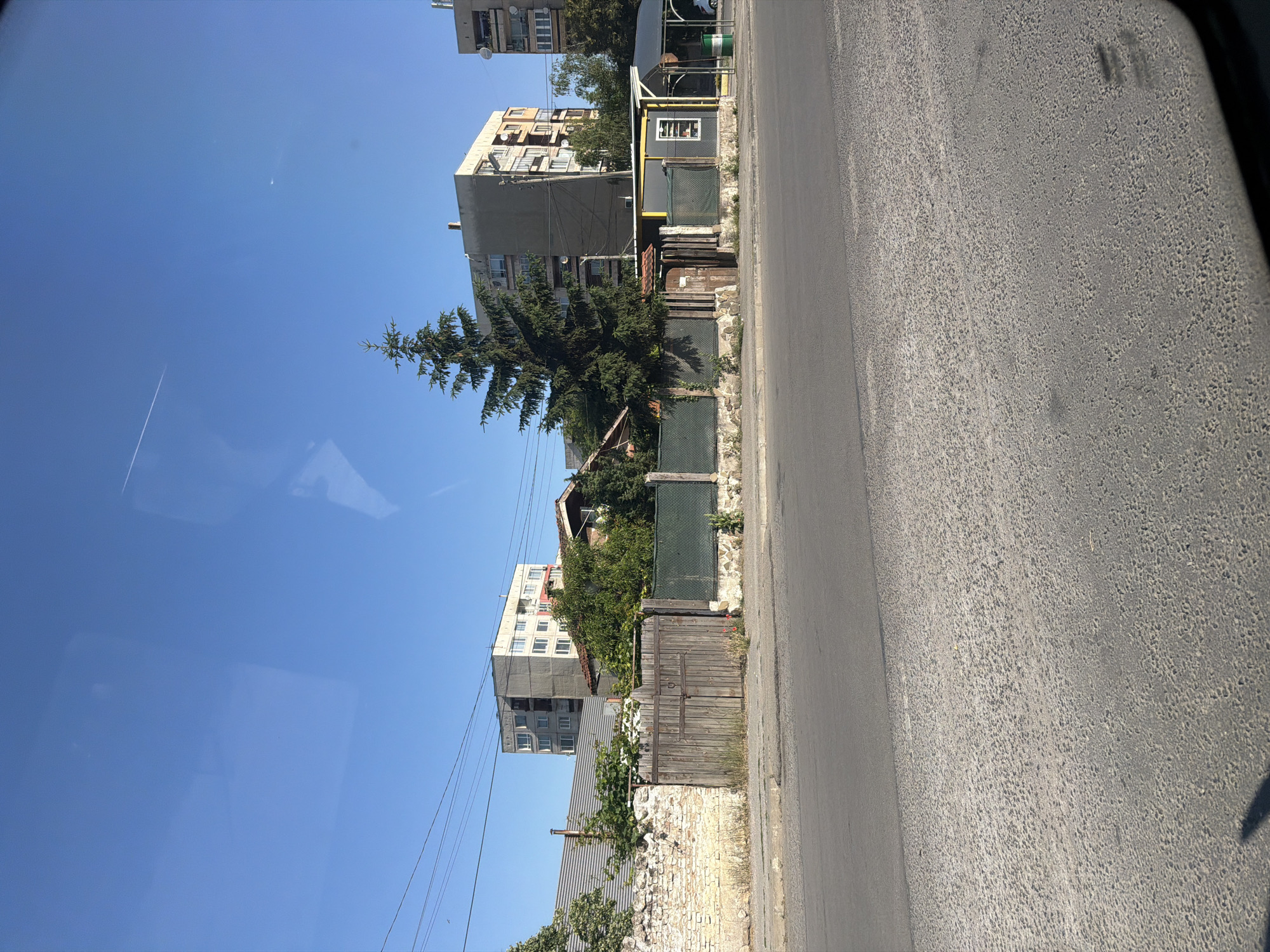 The image depicts a street scene in the city of Radomir, Bulgaria, on a clear, sunny day. In the foreground, a gray asphalt road with a rough texture is visible. Beyond the road, a low stone wall with some crumbling sections is present, bordering a property with a wooden gate and a green fence. Behind the fence, trees and vegetation are abundant. Several multi-story apartment buildings, typical of Soviet-era architecture, are visible in the background. A tall evergreen tree stands prominently in the mid-ground. The sky is a clear blue, with a faint airplane contrail visible. The overall impression is a residential area within a European city.