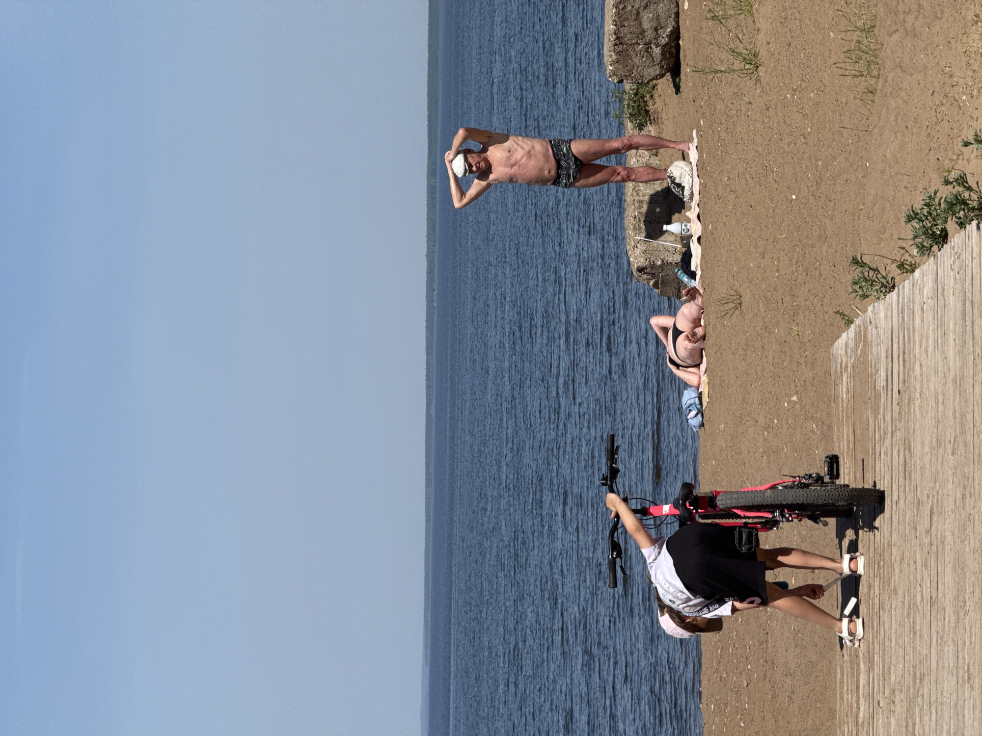 The image depicts a beach scene. In the foreground, a person is standing on a wooden boardwalk with a red bicycle. They are wearing a white shirt, black skirt, sandals, and a pink cap. Behind them is a body of water, with the opposite shore visible in the distance.
Further back on the beach, a woman in a black bikini is lounging on a pink towel. A man wearing patterned swim trunks and a white hat is standing nearby. He has his hands behind his head. A white towel and a water bottle are situated next to them. The environment includes sand, grass, and some rocks.
The location is a beach in the city of Unknown, Unknown.