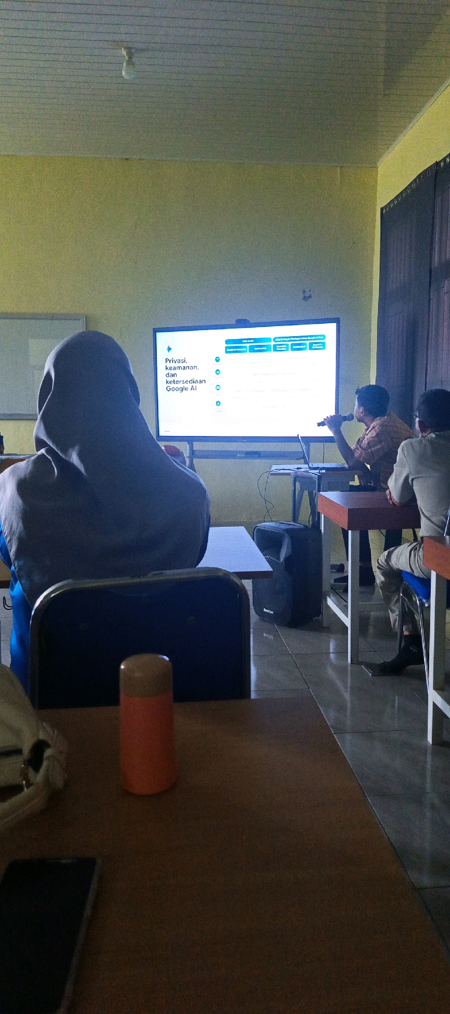In a classroom setting in Desa Sarang Mandi, Indonesia, a presentation is underway. A large screen displays information, with text visible in Indonesian. The title of the presentation appears to be "Privasi, keamanan, dan ketersediaan Google AI" (Privacy, security, and availability of Google AI). A presenter, an adult male wearing a patterned shirt, stands near the screen, holding a microphone and facing the audience. He is positioned to the right of the screen, with a laptop on a table in front of him. Two audience members are visible: one is a person from behind wearing a hijab, seated in the foreground and facing the screen, and another is a person seated to the right of the presenter, wearing a light-colored jacket and trousers. The room has yellow walls, a white ceiling with light fixtures, and tiled flooring. A dark speaker is positioned on the floor to the right of the presenter's table. In the foreground, a wooden table holds a smartphone and an orange cylindrical object. The overall scene suggests an educational or informational session in progress.