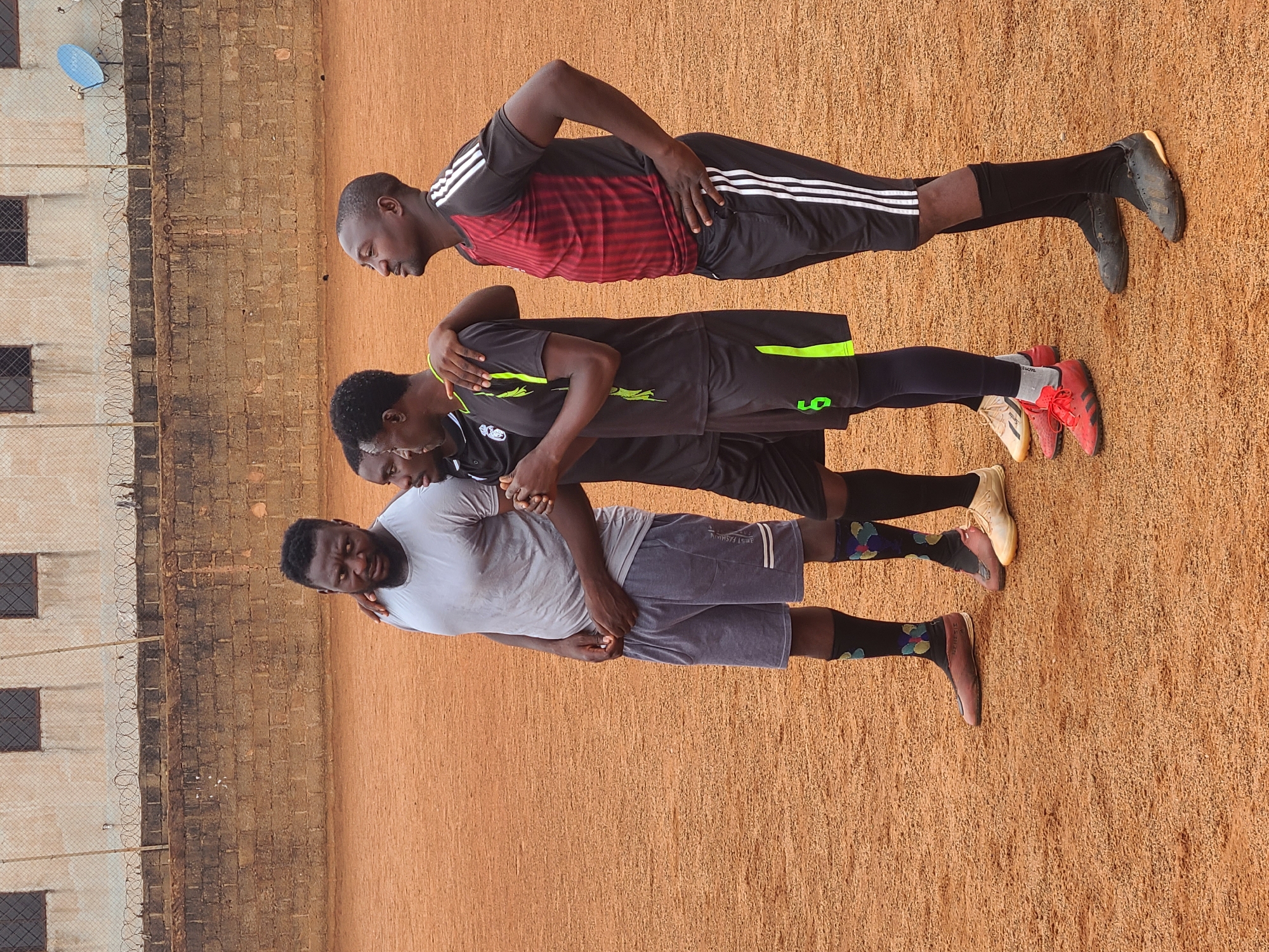 Four men stand in a line on a dirt surface, possibly a sports field, in the city of Unknown, Unknown. The backdrop features a brick wall topped with wire fencing and a building with windows.
The first man wears a black and red striped soccer jersey with white stripes on the shorts, black socks, and black soccer cleats. He has his hands on his hips.
The second man wears a black soccer jersey with a neon green stripe on the side, black shorts, black socks, and red soccer cleats. His arm is around the first man.
The third man wears a black jersey with a white logo and black shorts. He also has a hand on the second man.
The fourth man, at the front of the line, wears a gray T-shirt and gray shorts with white stripes. He has black socks with colorful decorations and brown shoes. He has his hand on the third man.
All four men face the same direction and appear to be posing or standing together.