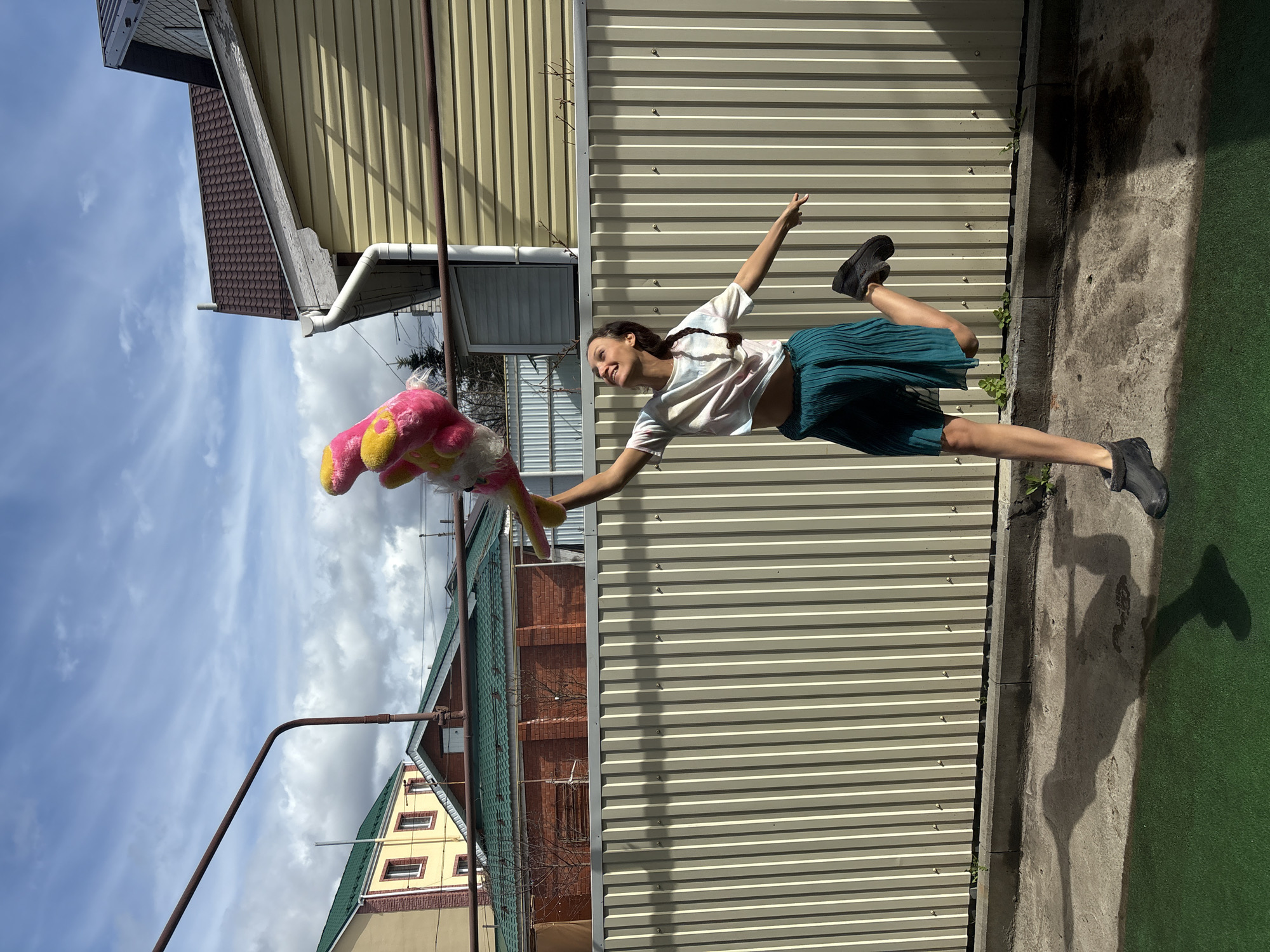 A woman is mid-leap in an outdoor setting. She holds a large, colorful plush toy. She wears a light-colored shirt, a dark green skirt, and black shoes. The background includes a corrugated metal fence, a building with siding and a tiled roof, and a sky with scattered clouds. She is positioned against the fence in the yard. The location is Ulyanovsk, Russia.