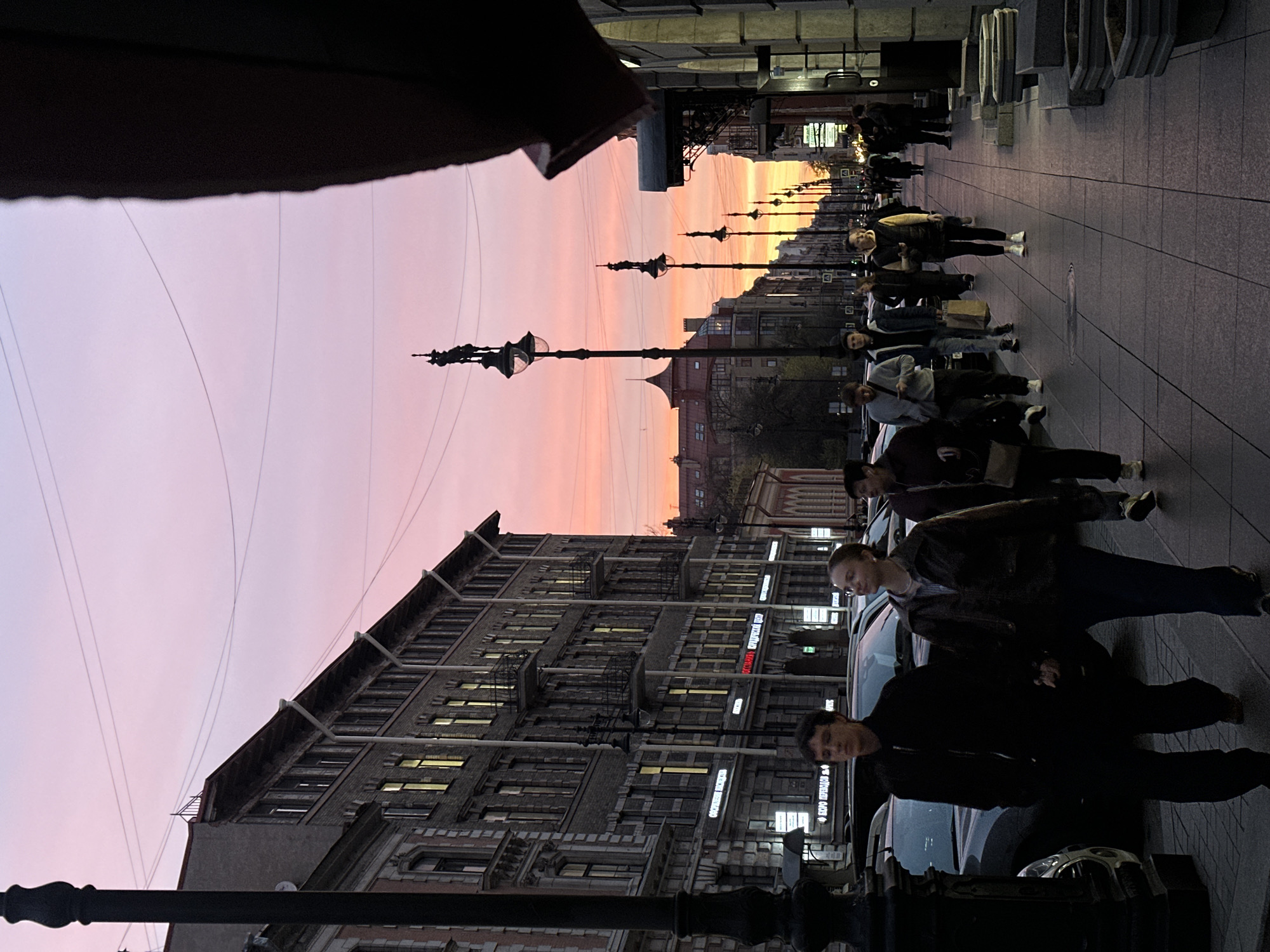 The image shows an urban streetscape at dusk, likely in an unidentified city. A row of buildings with lit windows stretches into the distance on the left side of the frame. The sky is a blend of pink and orange hues, suggestive of sunset or sunrise. Overhead wires run across the sky.
A line of people is walking along the sidewalk on the right. Street lamps are interspersed along the street, adding to the urban scene. Cars are parked along the curb, partially obscuring the facades of the buildings.