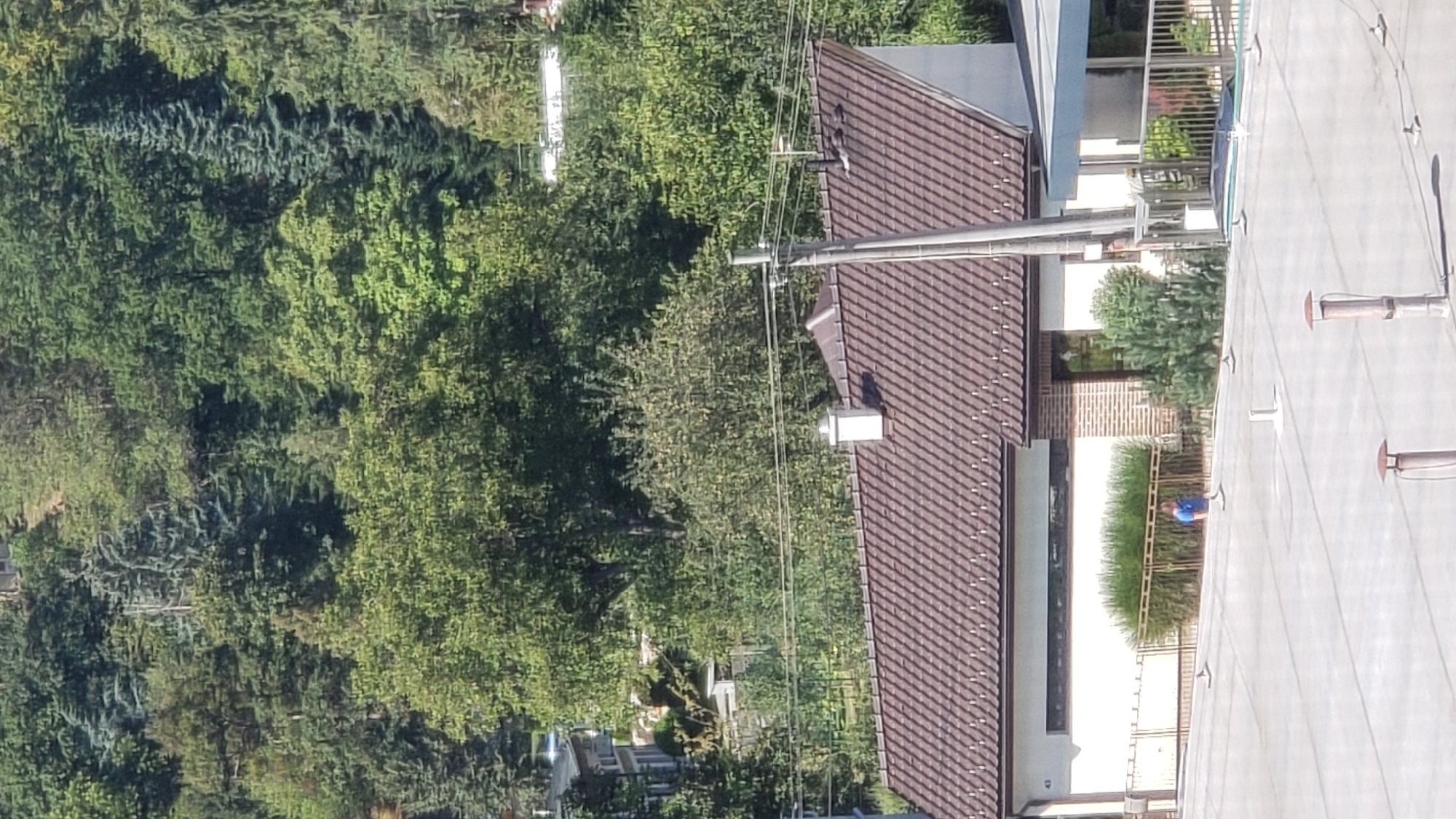 The image captures a view of a building with a brown tiled roof, a white facade, and a brick section. A person wearing a blue shirt is standing on a gray, flat rooftop in the foreground. The building is situated amidst lush green trees, suggesting a suburban or semi-rural setting. In the distance, more trees and vegetation dominate the landscape, with glimpses of other structures hinting at a developed area. Power lines and poles are visible, indicating the presence of infrastructure. The scene appears to be taken during daylight, under a clear sky. There is no specific identifiable landmarks visible that would pinpoint the exact location in Prešov, Slovakia, but the overall environment suggests a residential or commercial area with significant greenery.