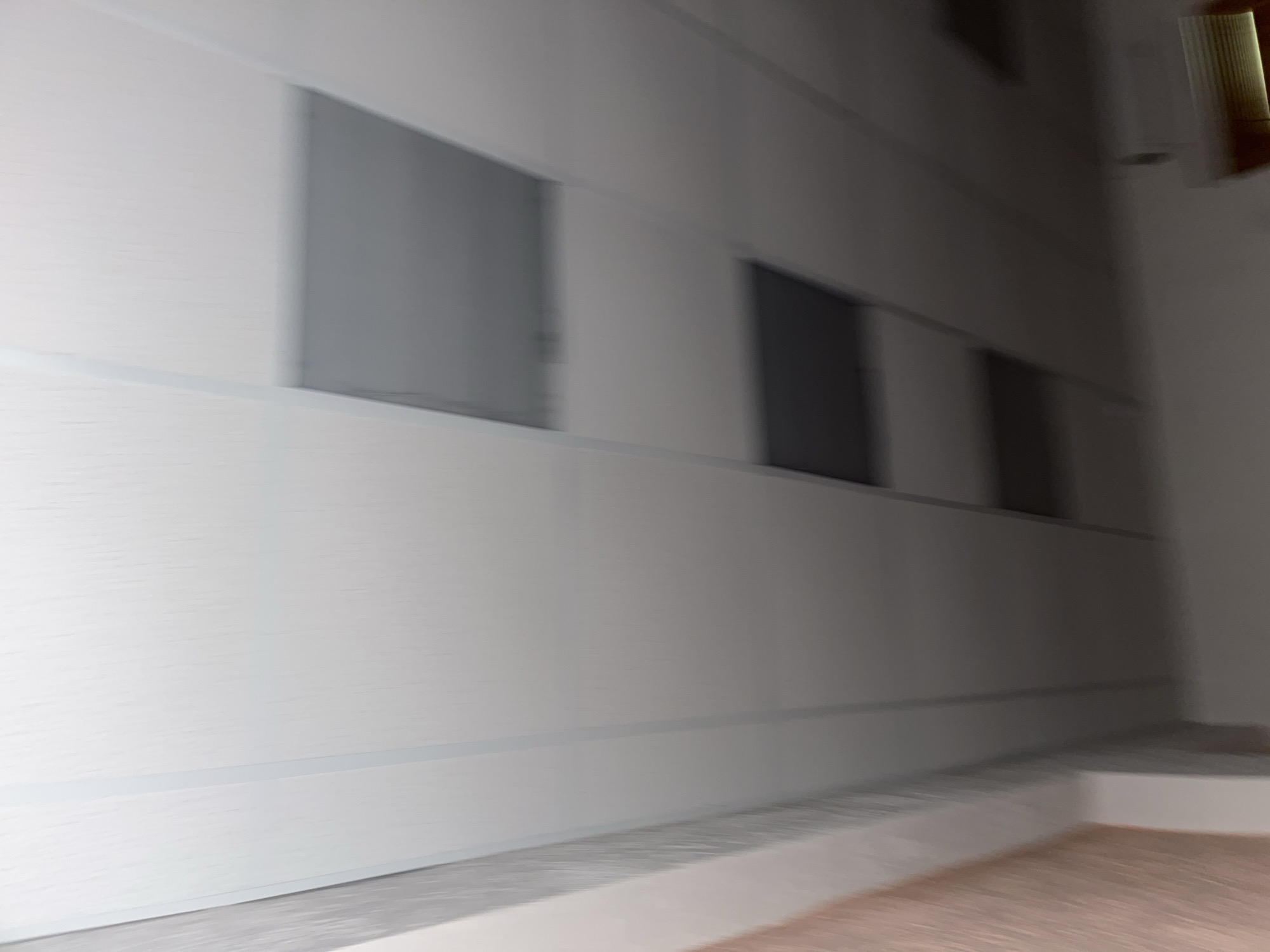 This is a medium-shot, blurry photograph taken from a low angle, looking upwards at a white wall with three dark, rectangular recesses. The recess on the left appears to be a window, while the other two appear to be doorways or deeper recesses. The wall itself has a textured, slightly horizontal grain. There is a darker horizontal line running across the wall about a third of the way up from the bottom, and another similar line above it. The floor in the foreground is a light grey, with a lighter band running horizontally across it, suggesting a step or a raised area. The right side of the image is darker, with a vertical shadow or recess visible. At the very top right of the image, there is a bright, vertical object with horizontal lines, possibly a piece of trim or a light fixture. The overall impression is of an interior space, possibly a hallway or corridor, with modern architectural features. The time of day and weather are not discernible. There is no visible text in the image. The blurriness suggests motion during the capture.