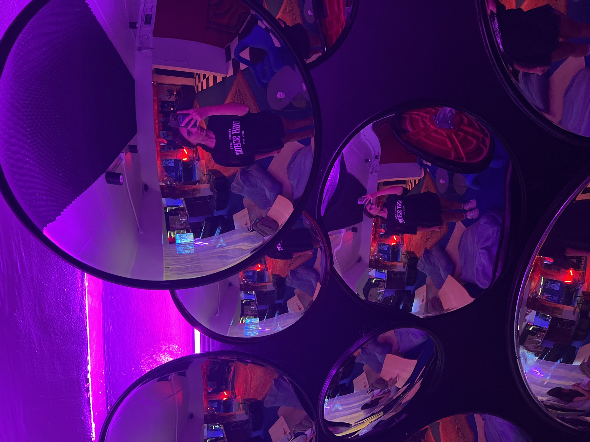 The image captures a person taking a selfie in front of a wall adorned with circular mirrors, which reflect the surrounding environment. The scene is illuminated with a prominent purple lighting, casting a vibrant hue over the entire setting. The reflections in the mirrors reveal a room with colorful decor, including tables and chairs, and possibly additional lighting fixtures. The person in the reflection appears to be wearing a black T-shirt. The overall ambiance suggests an indoor space with a contemporary or artistic design. The location is identified as Phuket City Municipality, Thailand.