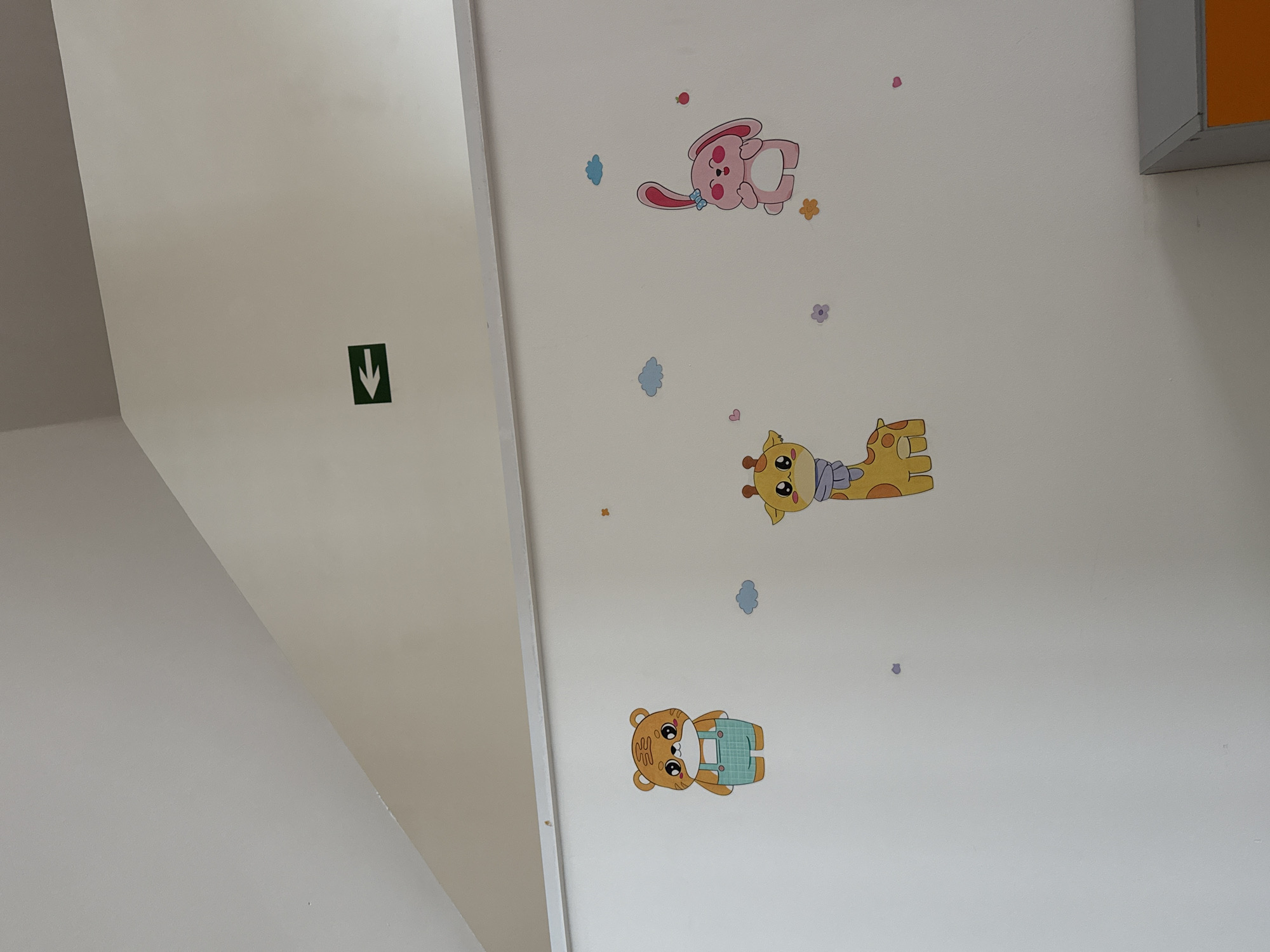 An indoor scene showcases a light-colored wall, likely white or off-white, decorated with a collection of whimsical cartoon animal decals. On the left side of the wall, a square green sign with a white downward-pointing arrow is prominently displayed, indicating a directional cue or emergency exit.
Three main animal characters are arranged vertically on the wall. At the top, a pink cartoon rabbit, adorned with a blue bow, is depicted lying on its back. Below it, a yellow cartoon giraffe, wearing a light blue scarf, is positioned. The lowest decal features a brown cartoon bear dressed in blue and white plaid overalls, also lying on its back. Surrounding these larger figures are smaller decorative stickers, including light blue cloud shapes, tiny flowers in various colors like pink, orange, and purple, and small red hearts, enhancing the cheerful and playful ambiance.
Architectural elements include an angular wall on the left, suggesting a corner or a section of a staircase. In the upper right corner, a small fragment of a fixture, possibly a cabinet or shelf, with an orange front and a grey side, is visible. The scene is brightly and evenly lit, typical of an indoor environment. There are no people present, and no specific activities are occurring. The only visible symbol is the white arrow on the green sign. This decorated interior space, located in Yerevan, Armenia, evokes a child-friendly atmosphere, possibly a daycare, school, or waiting room.