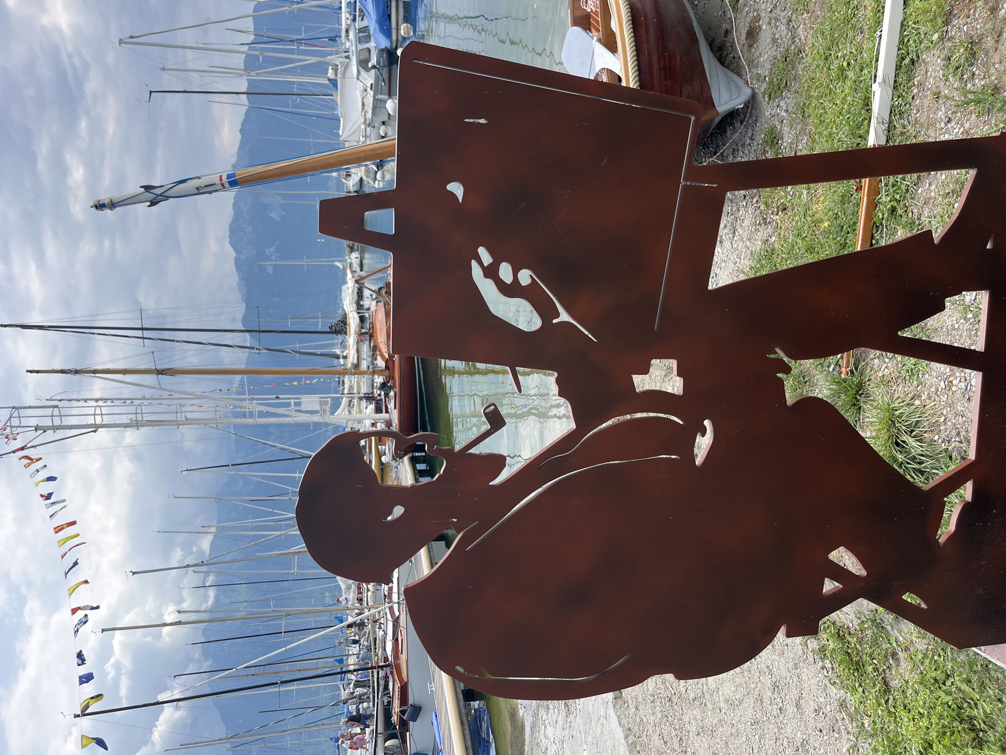 The image depicts a silhouette sculpture of an artist, likely in a seated position, holding a pipe and facing an easel. The sculpture is made of dark, reddish-brown metal. The easel displays a cutout of a hand holding a brush or pen, with a crescent moon shape. Behind the sculpture, a harbor is visible with numerous sailboats docked. The water of the lake is calm, and mountains form a backdrop. A string of colorful nautical flags is strung across poles in the upper portion of the image. The ground in front of the sculpture is a gravelly path with patches of green grass. The scene is located in the city of La Tour-de-Peilz, Switzerland.