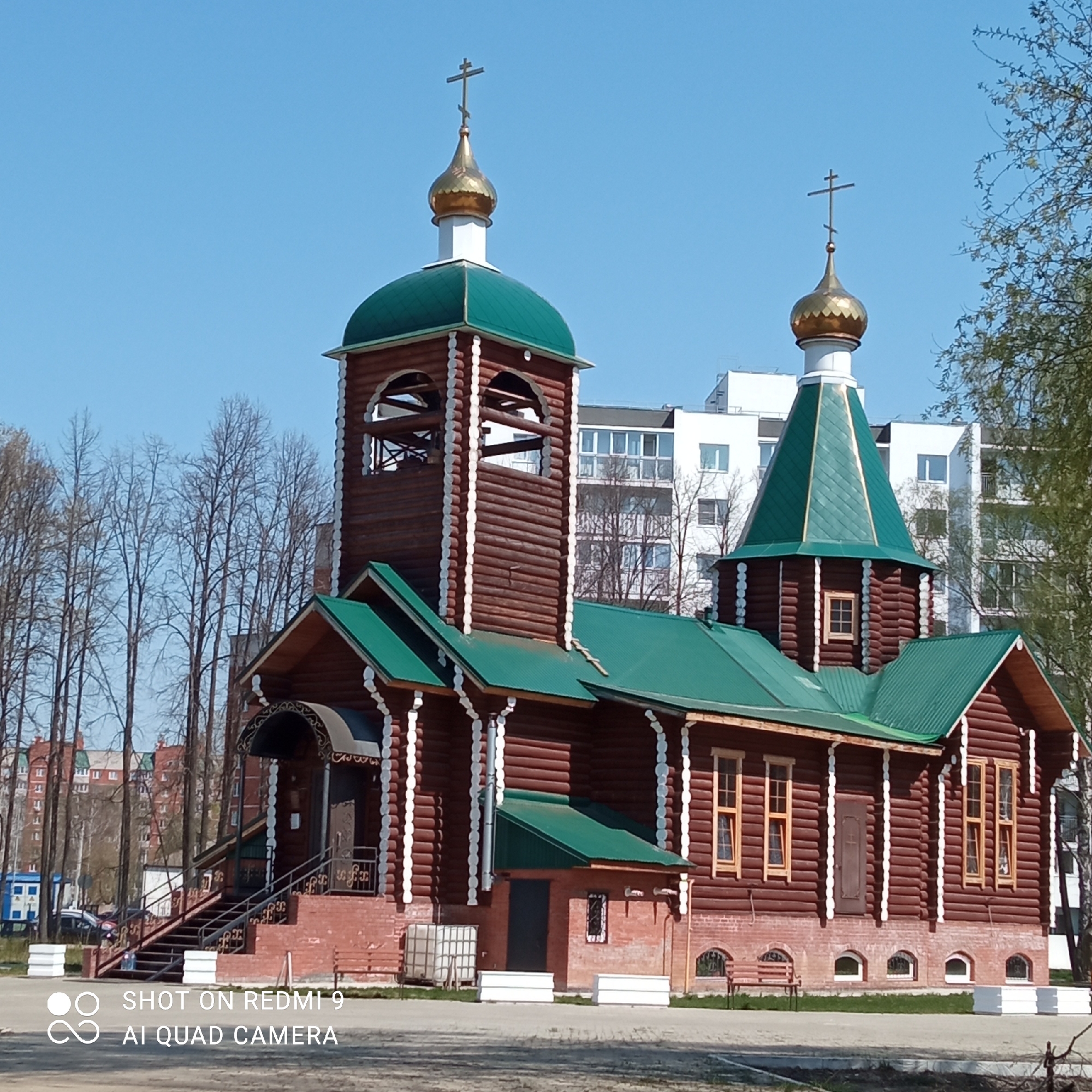 The image depicts a wooden Russian Orthodox church with green roofs and golden domes, located in Chaikovsky, Russia. The church has a bell tower and is constructed primarily of logs with white trim. In the background, there is an apartment building. The foreground shows a paved area. Trees surround the church. The sky is clear and blue. There are no people visible in the image.