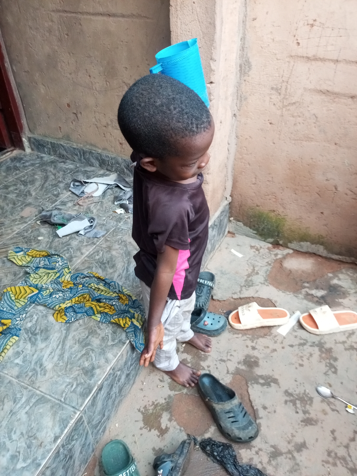 A young child stands on a concrete floor next to a tiled step in Tempure, Nigeria. The child, facing away from the camera, is wearing a dark t-shirt with pink accents and light-colored pants. Scattered on the floor and steps are various items including a brightly colored patterned cloth, several pairs of shoes and sandals, and some discarded fabric scraps. A bright blue plastic container is visible leaning against a textured wall behind the child. The environment appears to be an outdoor area, possibly a courtyard or entrance to a building.