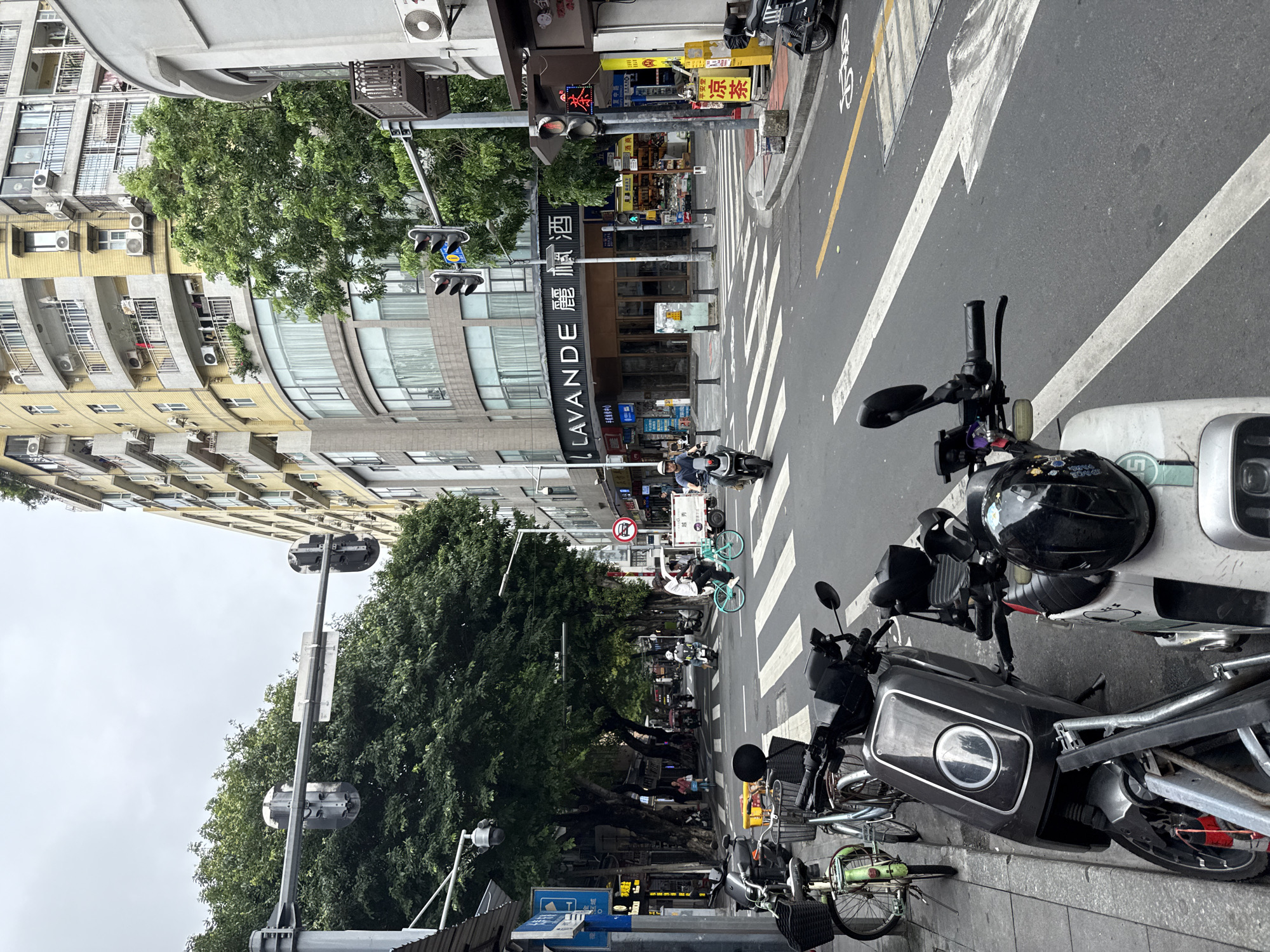 In Liwan District, China, a street scene unfolds under an overcast sky. The foreground features several parked electric scooters and bicycles on a concrete sidewalk, with a black scooter prominently displayed. Beyond the sidewalk, a wide asphalt street with crosswalk markings stretches into the distance. Several traffic lights are visible, with one set showing a red light. Pedestrians are seen crossing the street, and individuals are riding bicycles and scooters. Large trees with dense green foliage line the street, creating a shaded canopy. Buildings of varying architectural styles, including a tall, yellow residential building with numerous balconies and air conditioning units, are situated along the street. A store with a prominent sign reading "LAVANDE" is visible. The overall atmosphere suggests a busy urban environment during daytime.