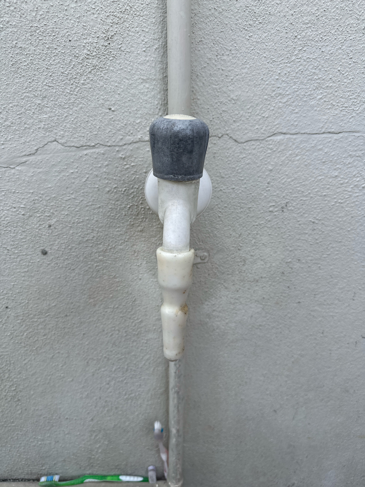 The image depicts a close-up, vertical view of an outdoor water faucet mounted on a textured, light-colored wall. The faucet is made of white plastic with a dark gray rubber knob. A thin, light-colored pipe runs vertically behind the faucet, attached to the wall by a white plastic bracket. Below the faucet, a section of the same white plastic pipe extends downwards. At the very bottom of the frame, on the ground, lie two toothbrushes; one is green and white, and the other is a smaller, gray and white one. The wall itself shows some minor cracking and has a rough, pebbled texture. The scene appears to be located in Agara, India.