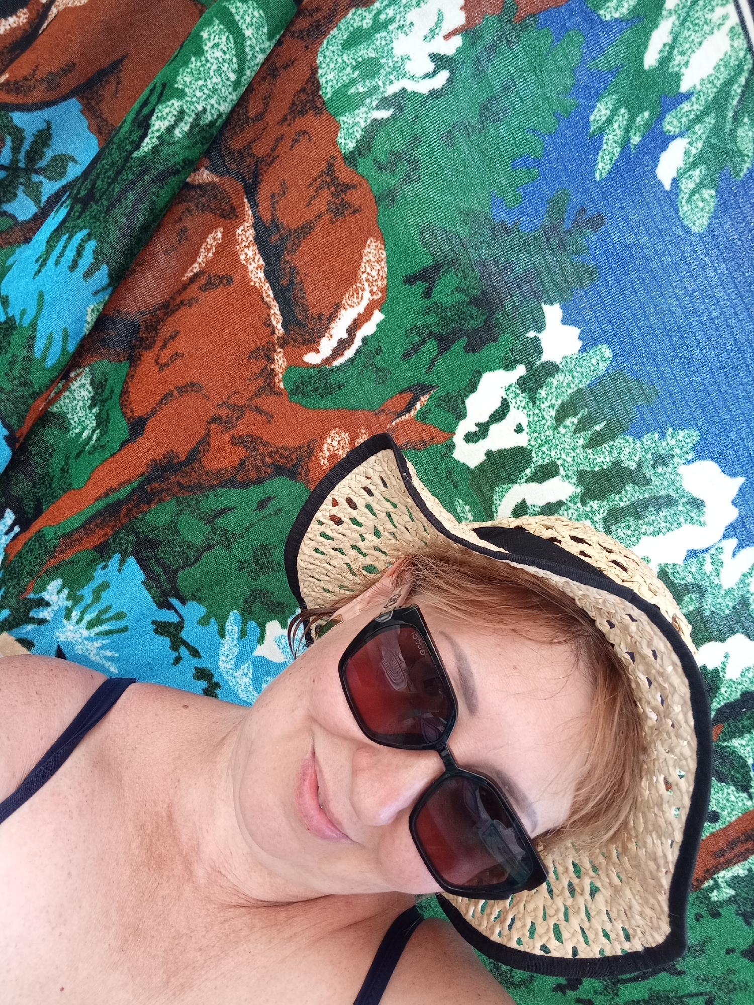 A woman with fair skin and red hair is lying down on a colorful, patterned fabric. She is wearing a straw hat with a black band and large, dark sunglasses. She is also wearing a dark-colored swimsuit. The fabric appears to be a beach towel or sarong with a large, abstract print of brown, green, blue, and white shapes. The woman is smiling and looking up at the camera. The image is a selfie taken from a slightly elevated angle. The scene does not provide any specific details to indicate a location within Озёрки ауылдық округі, Kazakhstan.