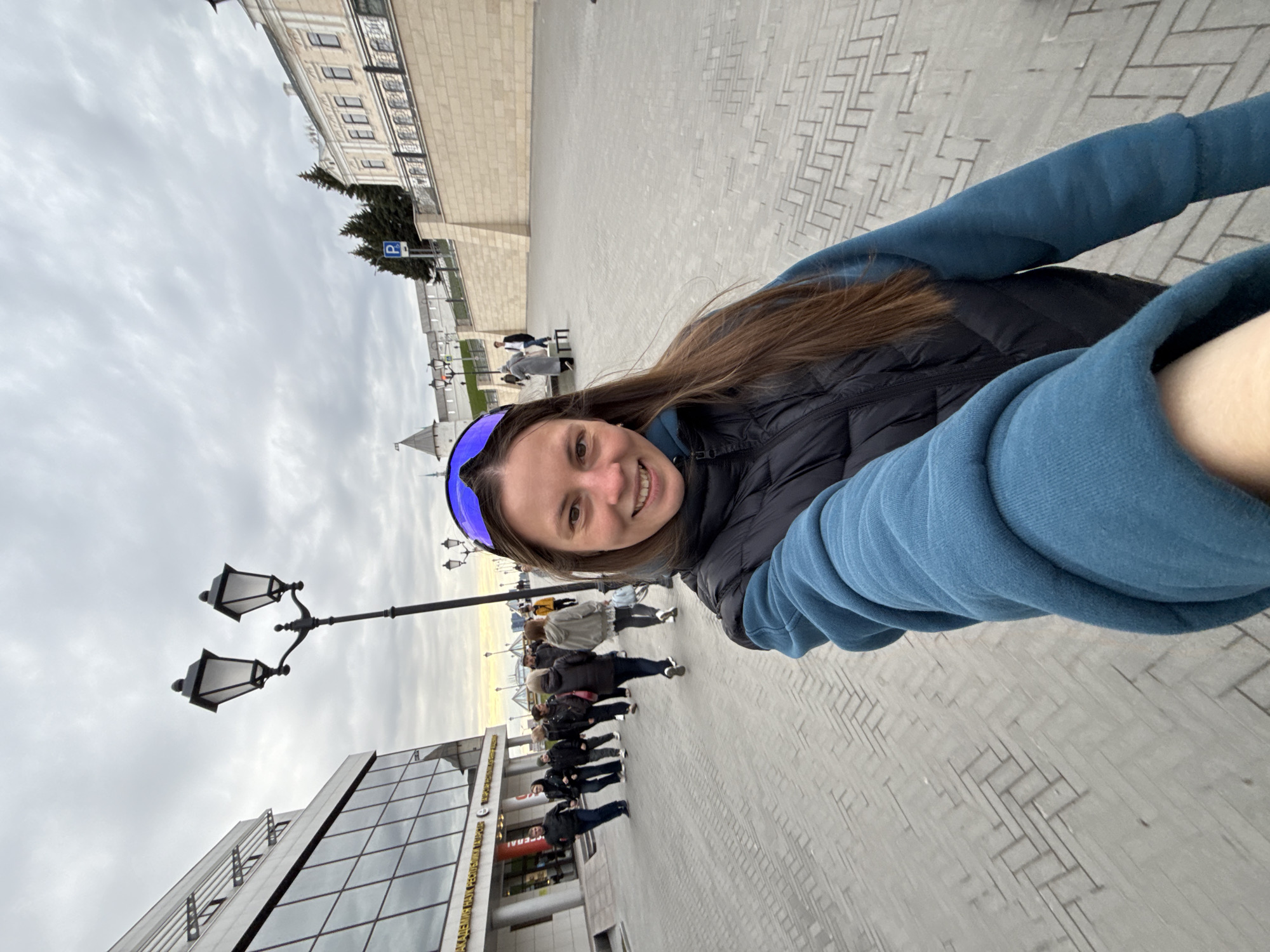 The image is a rotated selfie, depicting a smiling young woman in the foreground looking directly at the camera. She has long brown hair, is wearing a black puffer vest over a blue long-sleeved shirt, and a blue visor or athletic sunglasses pushed up on her forehead. Her left arm is extended towards the camera, holding the device.
The background shows an active urban street or square, paved with light-colored, patterned bricks. Numerous pedestrians are visible in the middle ground, walking in various directions. To the left, a tall, black, ornate street lamp with two lanterns stands. Behind it, a modern building features large glass windows, and Cyrillic text is partially visible on its facade, including "ФЕДЕРАЛ" (Federal) and "ТАТАРСТАН" (Tatarstan).
Further in the distance, on the right, are older, grand-looking buildings with white or light-colored stone facades. A distinctive white, pointed tower or spire is visible above these structures. Green trees and a blue parking sign with a white 'P' are also discernible. The sky is overcast, suggesting a cloudy day, yet there is ample daylight. The overall scene conveys a lively city atmosphere. Given the textual clues and context, the location is Kazan, Russia.