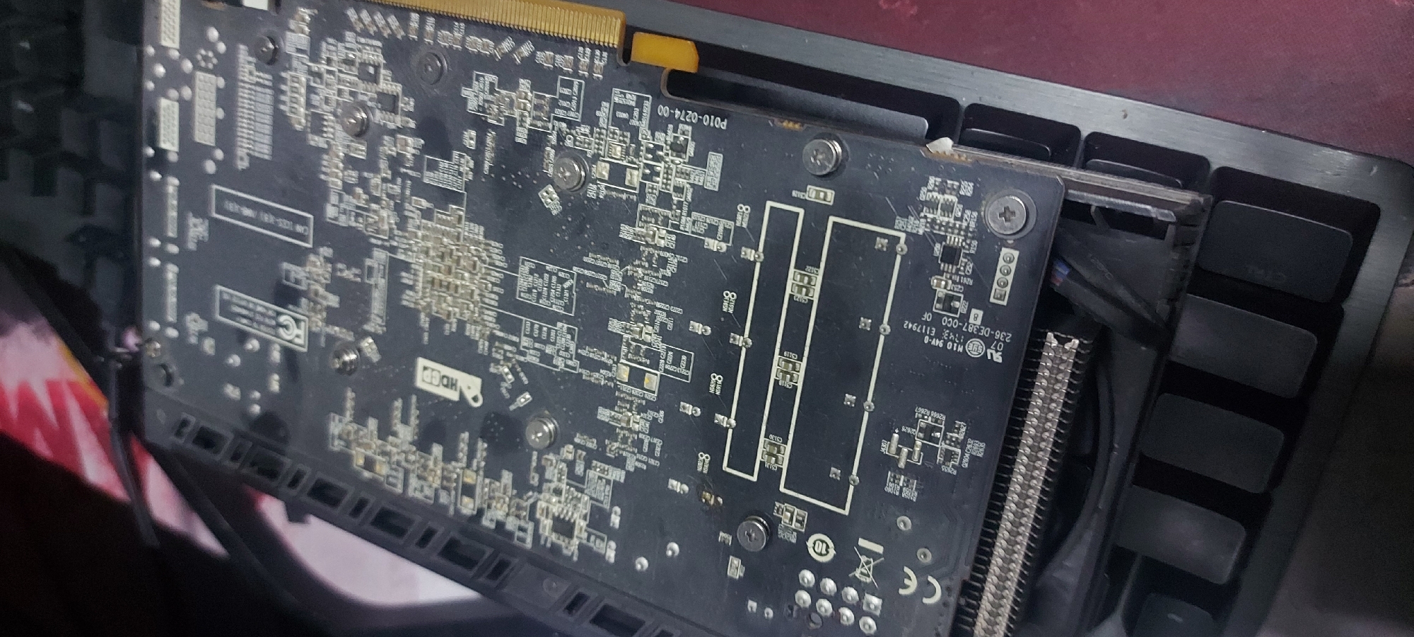 The image shows the back of a graphics card, likely used in a desktop computer. The printed circuit board (PCB) is dark gray or black, and is densely populated with surface-mount components. Several screws are visible, securing the PCB to the heatsink and backplate of the card. The PCI-e connector is visible at the top, with gold-colored contacts. Some text is visible on the PCB, including model numbers and regulatory markings like "CE" and "FCC". There's no evidence in the picture that would link this to Tashkent, Uzbekistan. It is impossible to infer a location from this image.