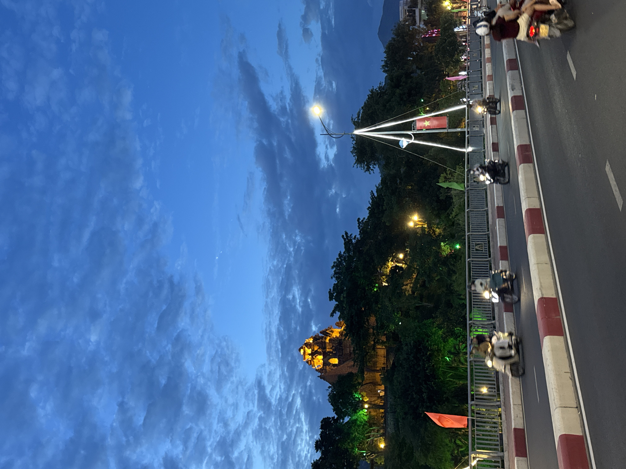 This image depicts a twilight scene in Nha Trang, Vietnam. A prominent feature is the Po Nagar Cham Towers, a historical complex illuminated with orange lights, situated on a lush, green hillside. The towers are surrounded by dense foliage and trees, with some areas subtly lit in green. In the foreground, a road with clear white and red demarcating lines carries vehicular traffic, primarily motorcycles. Several motorcycles are visible, some carrying multiple passengers. A bridge or elevated road is present, lined with a metal railing and decorated with lights. A tall, illuminated structure resembling a stylized sail or kite, adorned with white lights and a Vietnamese flag, stands near the bridge. The sky above is a deep blue, characterized by dramatic, textured clouds that suggest the transition from day to night. The overall atmosphere is dynamic, with the movement of the motorcycles and the ambient lighting contributing to a lively urban environment.
