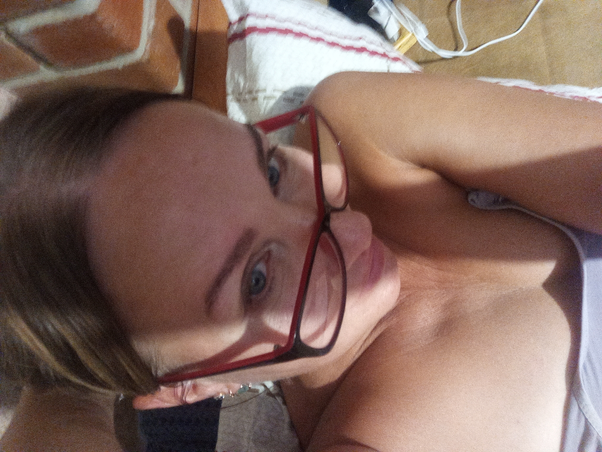 A young woman wearing red-rimmed glasses is lying down, looking up at the camera. Her head is tilted, and her eyes are partially visible through the lenses. She appears to be in an indoor setting, with a brick wall and a striped cloth visible in the background. A white cord is also present in the frame. The image is taken from a low angle, with the woman's face and upper body occupying most of the frame.