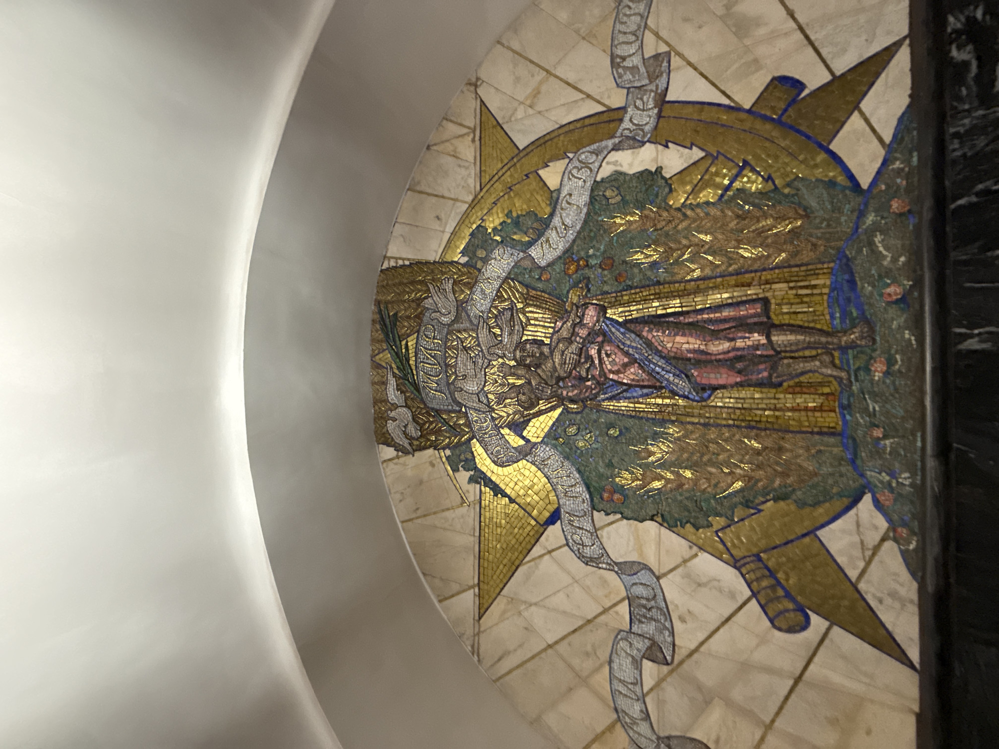 The image shows a mosaic artwork on an arched wall. The mosaic depicts a central figure holding a sickle and standing in a field of wheat. Behind the figure, a large golden star is prominent. Ribbons with Cyrillic text are draped across the mosaic. The artwork is elaborate and detailed, with a mix of gold, blue, green, and red tiles. The mosaic is set against a marble-like background. The environment appears to be indoors, possibly a public space like a metro station, given the arched ceiling and the style of the artwork which is reminiscent of Soviet-era public art. The bottom of the image shows a dark, possibly stone or marble ledge. The overall impression is one of grandeur and symbolic representation.