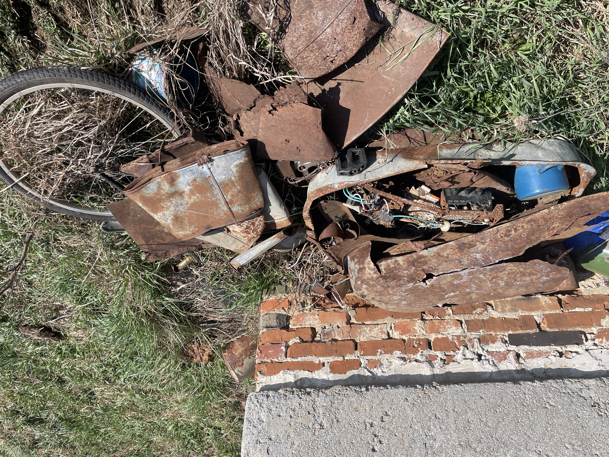 The image shows a patch of overgrown grass in what may be Поедуги, Russia, near a partially collapsed brick wall and concrete base. The scene is littered with rusted metal debris, including what appears to be parts of a bicycle wheel, a metal bucket, and fragmented metal sheets and containers. Electrical wires and components are also visible amidst the debris. The overall impression is one of neglect and decay.