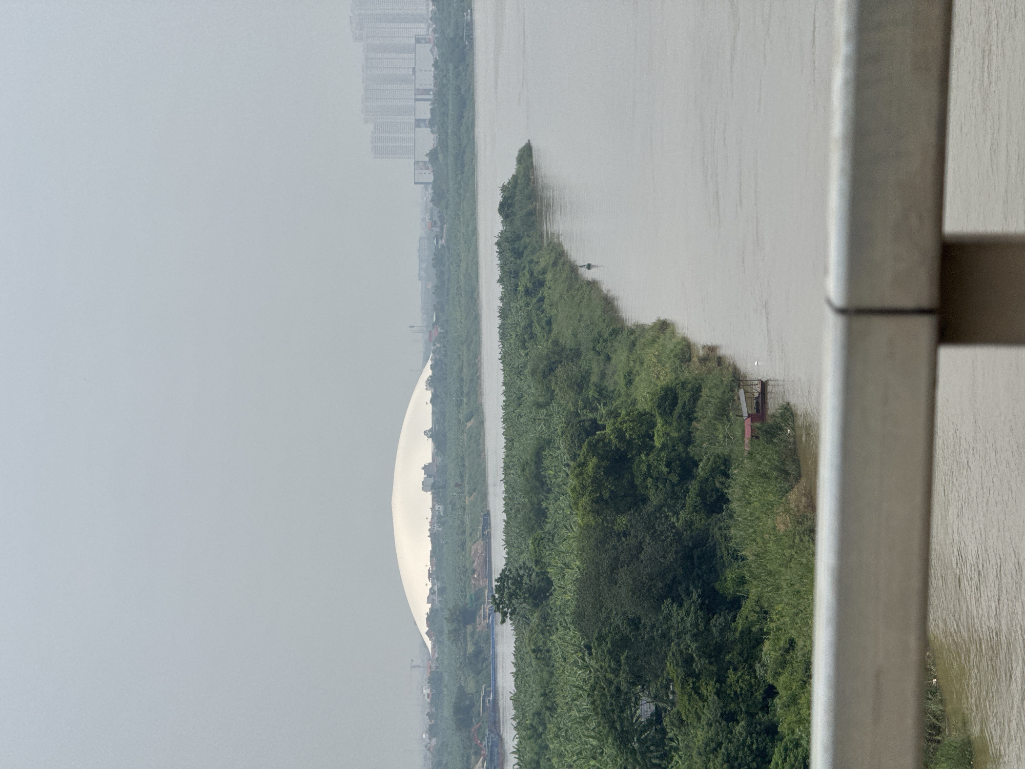 This is an outdoor, eye-level, vertical-format, daytime photograph taken from a bridge overlooking a wide, murky river in Hà Nội, Vietnam. In the middle ground, a dense, green, tree-covered island juts out into the river. On the island, there is a small, red, boat-like structure with a roof. Further in the distance, on the far bank of the river, a large, white, dome-shaped building is visible, with a city skyline featuring tall, modern buildings and some smaller structures in the background. The sky is overcast and hazy. The foreground is dominated by the railing of the bridge the photograph was taken from, which is a metallic gray color and slightly out of focus. There are no people visible in the image. The overall scene depicts an urban landscape with a significant natural element, the river and its vegetated island.