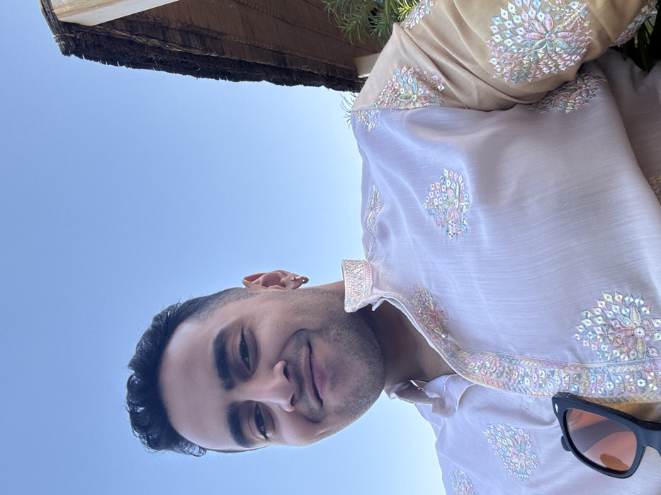 In the city of Malasar, India, a direct portrait shows an adult male from the chest up under a clear blue sky. The subject, positioned slightly left of center, has short, dark hair and a light stubble, looking at the camera with a subtle, closed-lip smile. A gold hoop earring is visible in his left earlobe. He is wearing a pale pink or lavender kurta featuring elaborate pastel-colored embroidery on the standing collar, central placket, and chest. The sleeves of the kurta are a contrasting golden-beige color, also detailed with matching embroidery. A pair of black sunglasses with orange-tinted lenses hangs from the placket on his left side. The background consists primarily of a bright blue sky. To the far right, a section of a building is visible, featuring a thatched roof composed of dried vegetation and a light-colored supporting structure, partially obscured by green foliage.