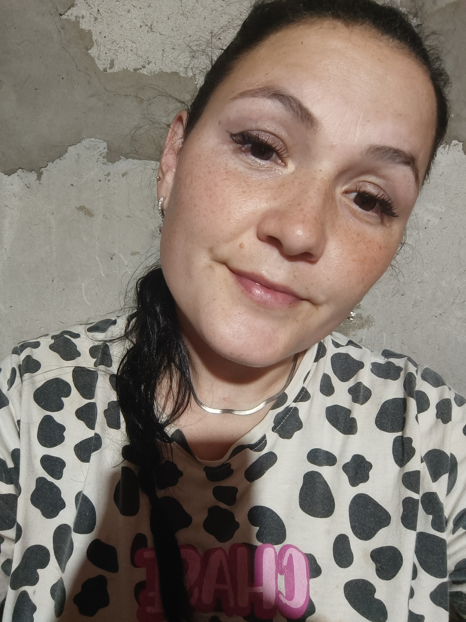 A woman with dark hair pulled back into a ponytail, wearing a black and white cow-print t-shirt and a thin silver necklace, looks towards the camera. She has light skin with freckles, dark eyebrows, and dark eyes. The background is a rough, grey concrete wall with some textured patches and cracks. The lighting is direct, casting some shadows and highlighting the woman's face. There is no indication of the city of Karsha, Russia, or any specific location beyond the described setting.