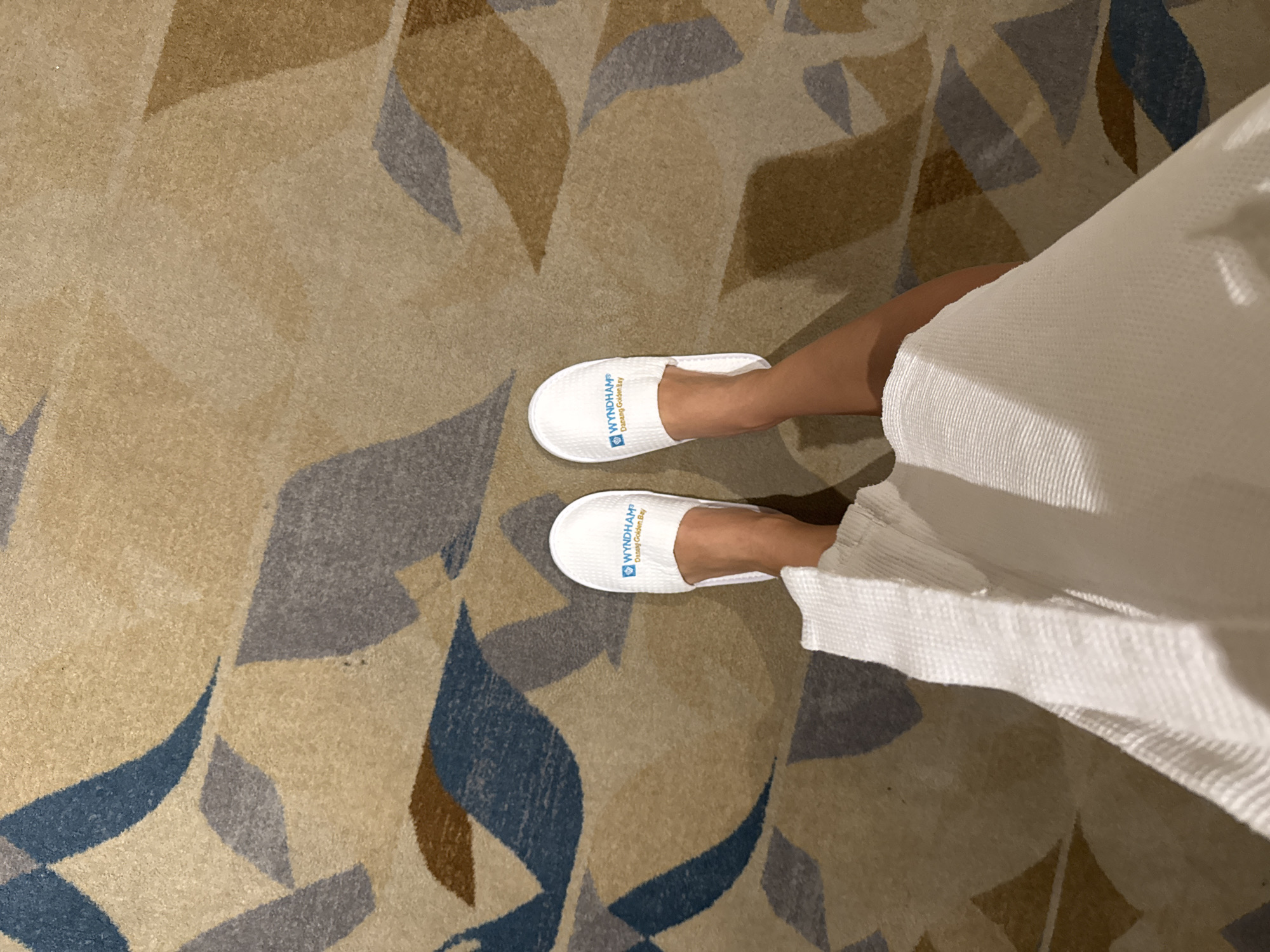 The image shows a person wearing a white bathrobe and white slippers standing on a patterned carpet. The slippers bear the logo "WYNDHAM® Danang Golden Bay". The carpet has a repeating leaf pattern in shades of brown, beige, gray, and blue. The location is Đà Nẵng, Vietnam.